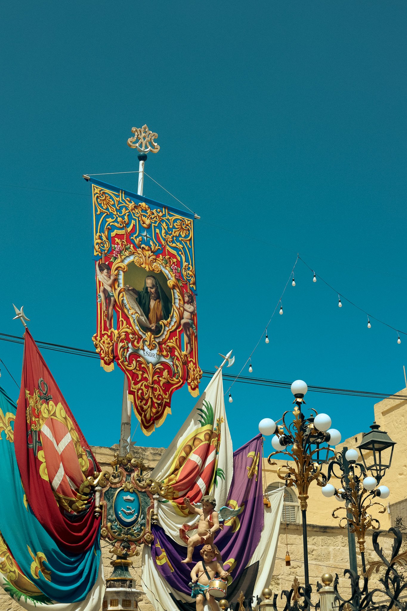 Decorative flags and banners with religious and heraldic symbols, including cherubs and a painting of a saint, displayed outdoors against a clear blue sky with street lamps and string lights.