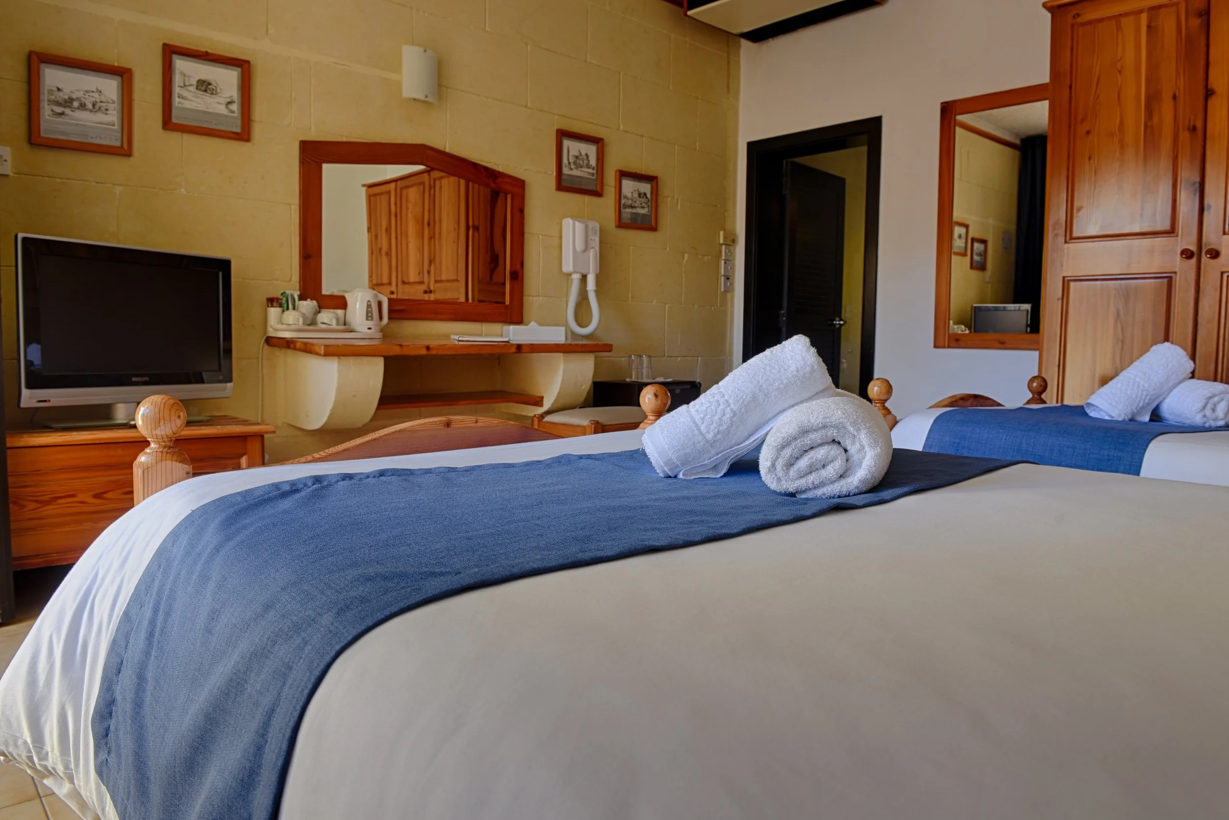 A hotel room with two beds, each with white towels rolled on blue bed runners, a small television on a wooden stand, framed pictures on the beige wall, a mirror, a desk with a mirror, a wall-mounted telephone, and a doorway leading to another room