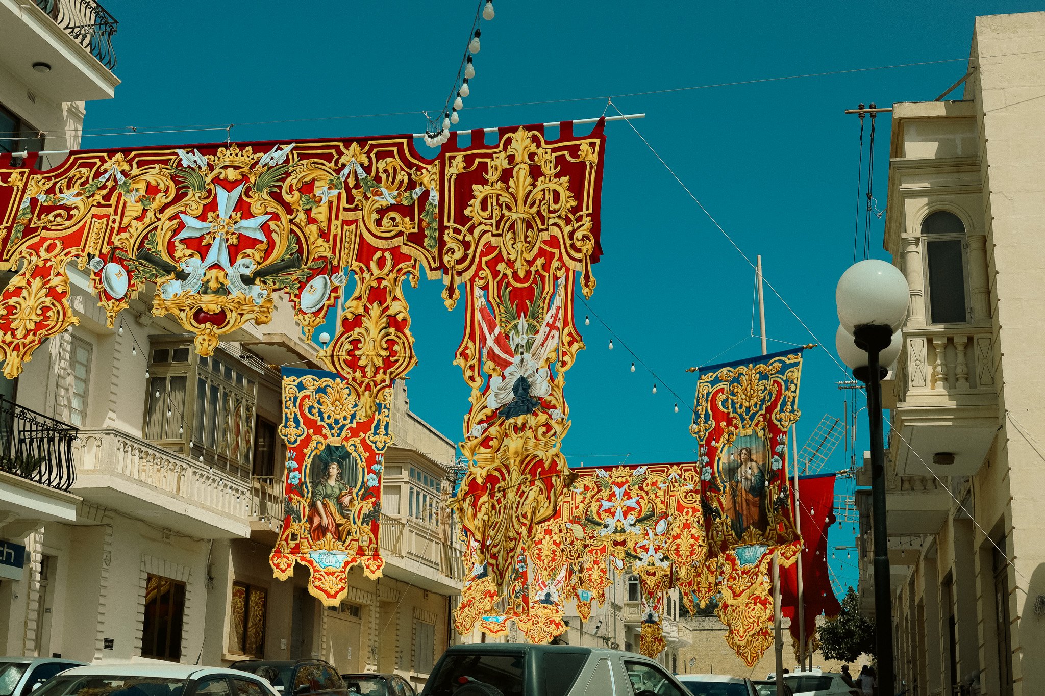 Colorful, ornate hanging banners decorated with gold, red, and white patterns, including religious imagery, hanging across a street lined with buildings, under a clear blue sky.