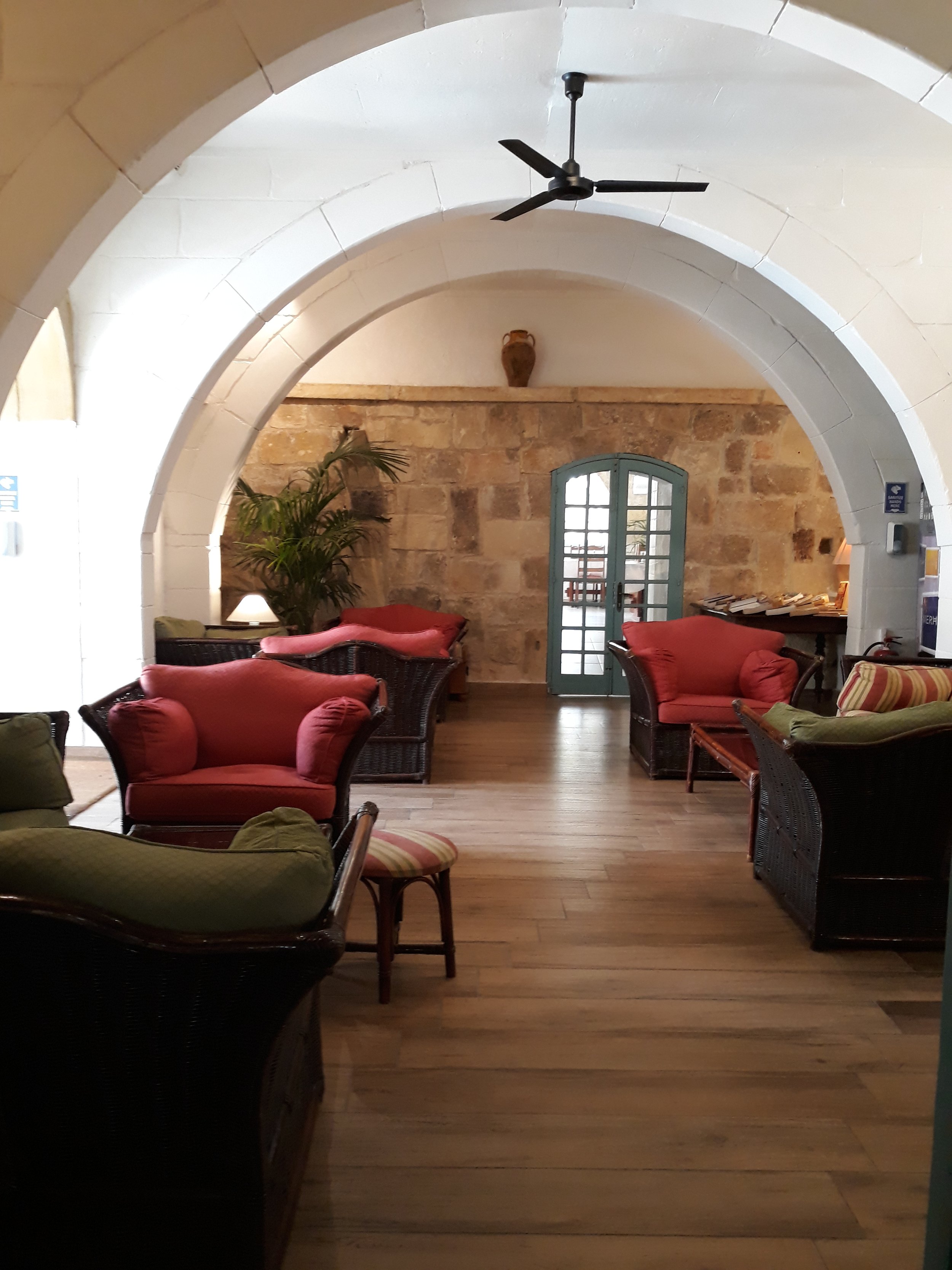 Interior of a lounge area with wooden floors, red and green cushioned chairs, wicker furniture, a potted plant, stone walls, arched doorways, and a ceiling fan.