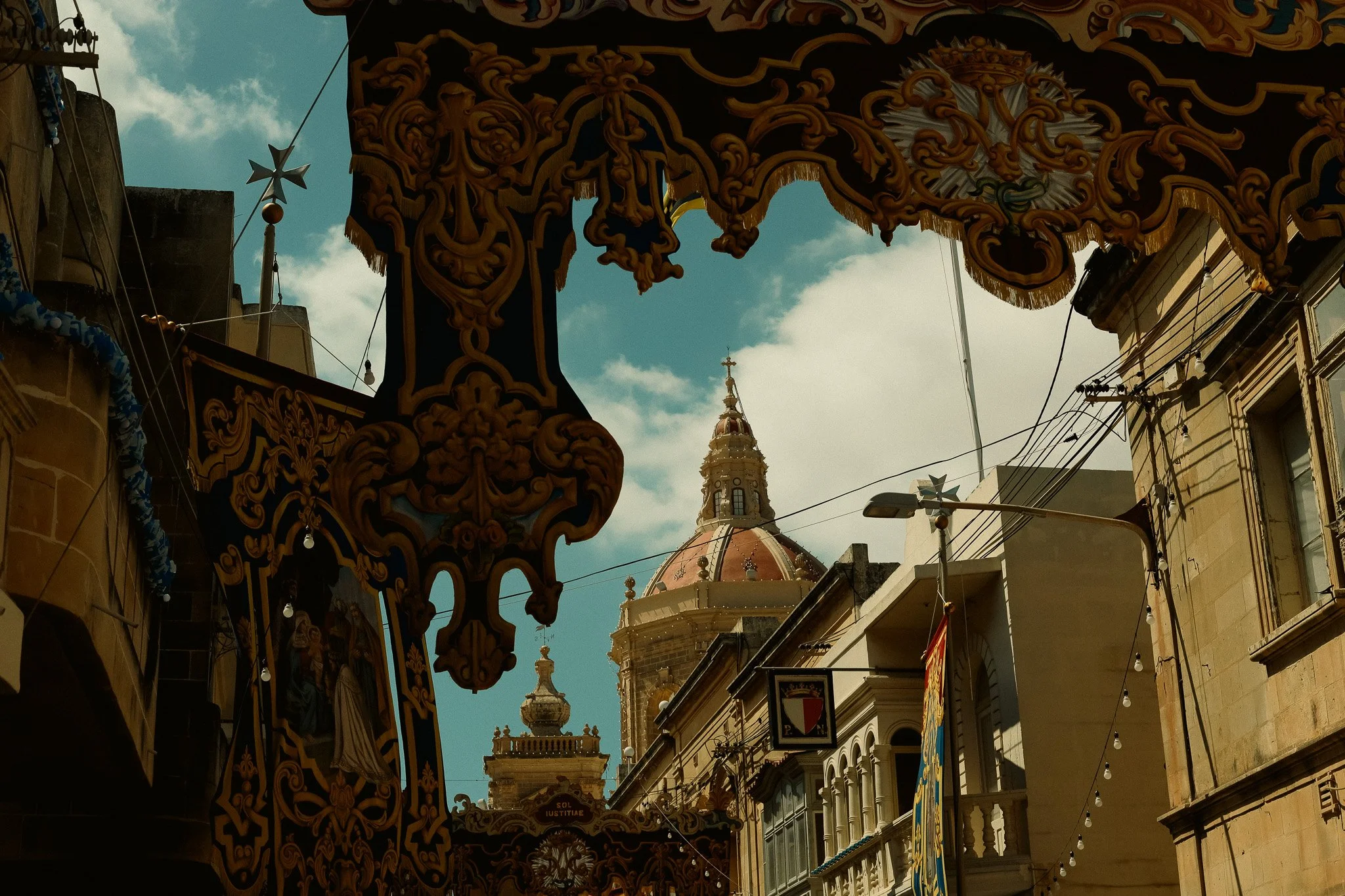 View of historic buildings and a church dome in a sunny street, framed by ornate decorations hanging overhead.
