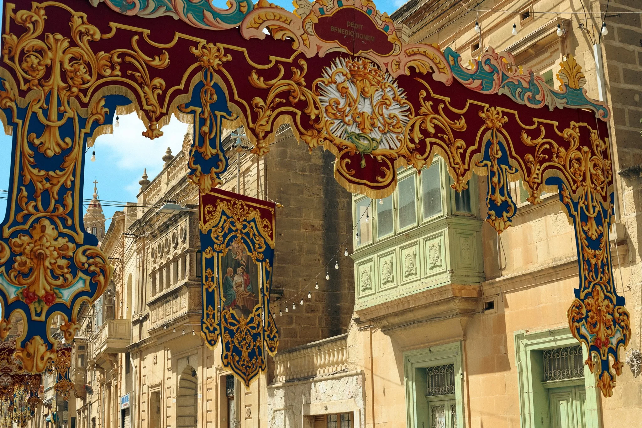 Decorative colorful fabric banners with intricate gold embroidery hanging in front of historic buildings with balconies and ornate window frames.