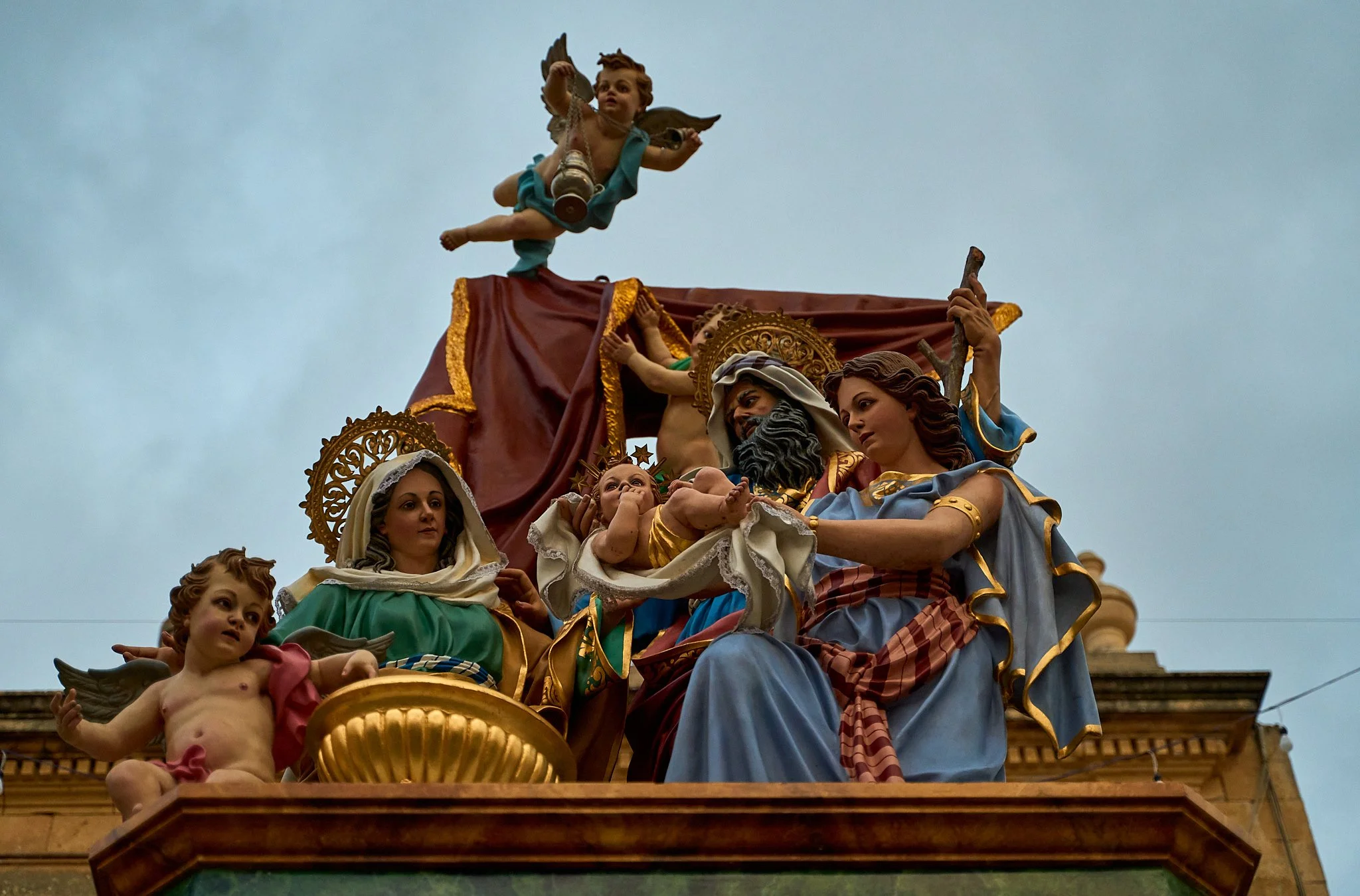 Colorful religious sculpture of the Virgin Mary, baby Jesus, and other Biblical figures on a rooftop, with a cloudy sky background.