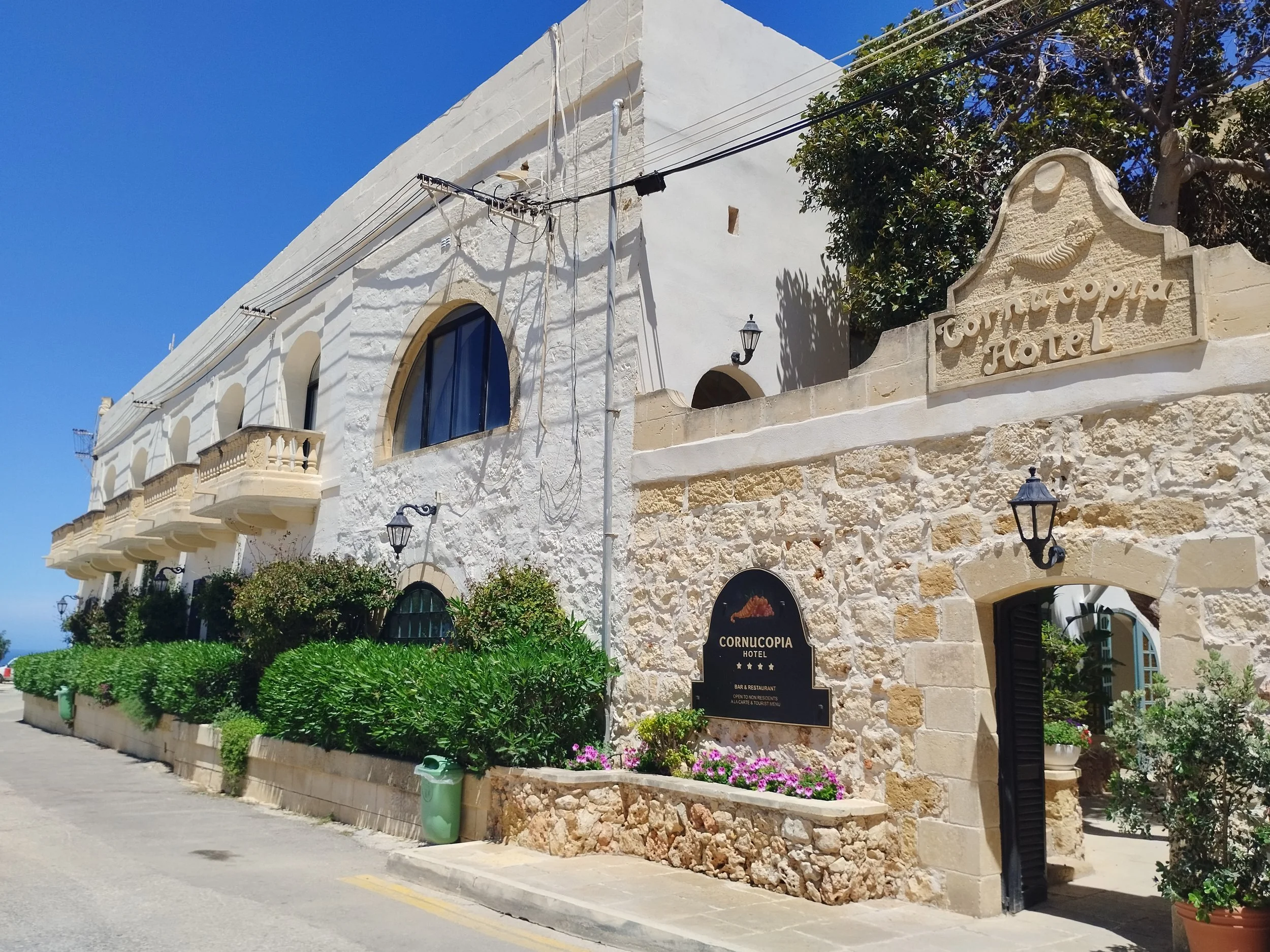 Exterior view of the Cornucopia Hotel, a historic stone building with arched windows, balconies, and decorative signs, surrounded by greenery and flower beds under a clear blue sky.