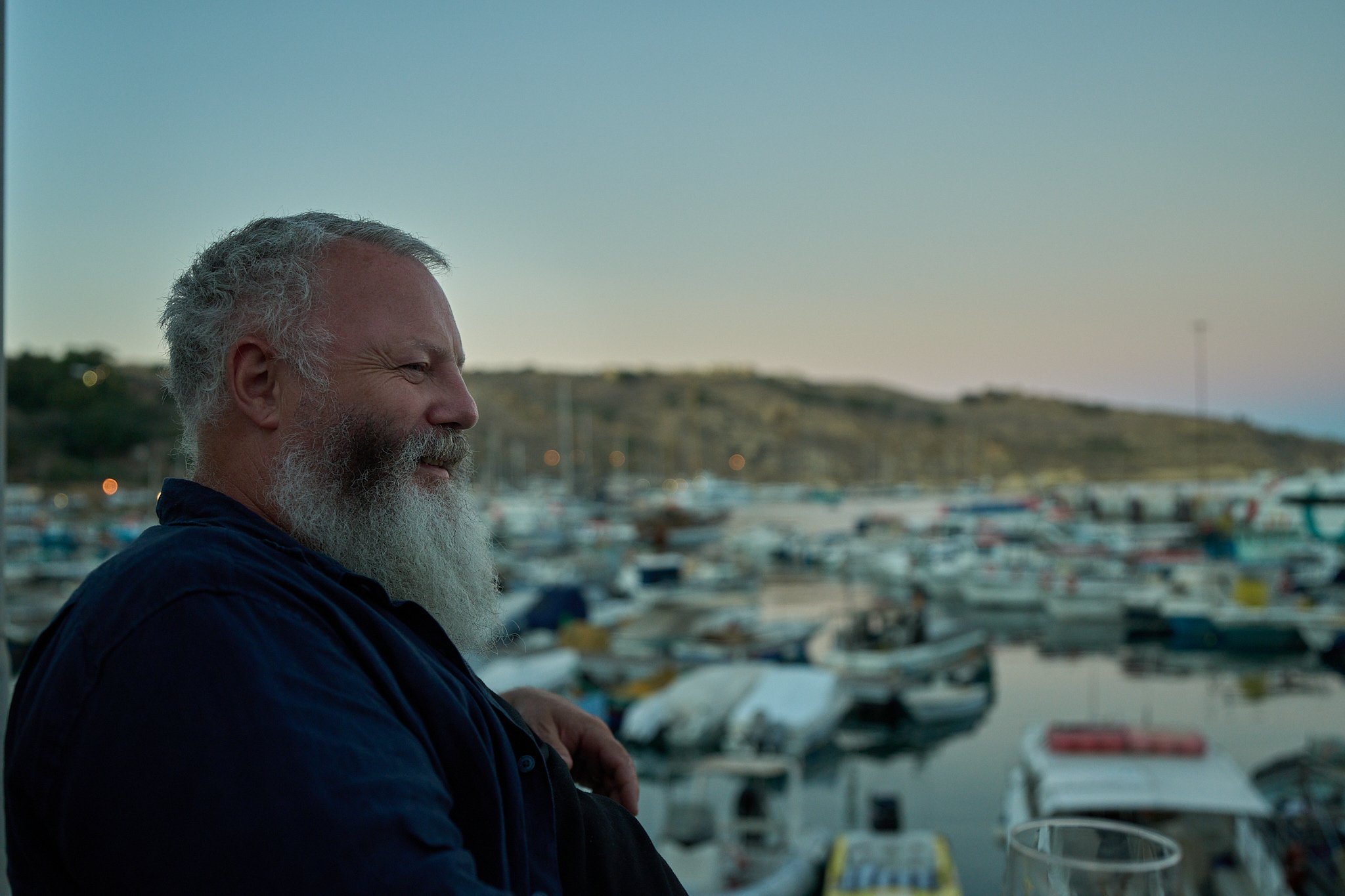 Profile of a smiling older man with a white beard and gray hair, overlooking a marina filled with boats at sunset.