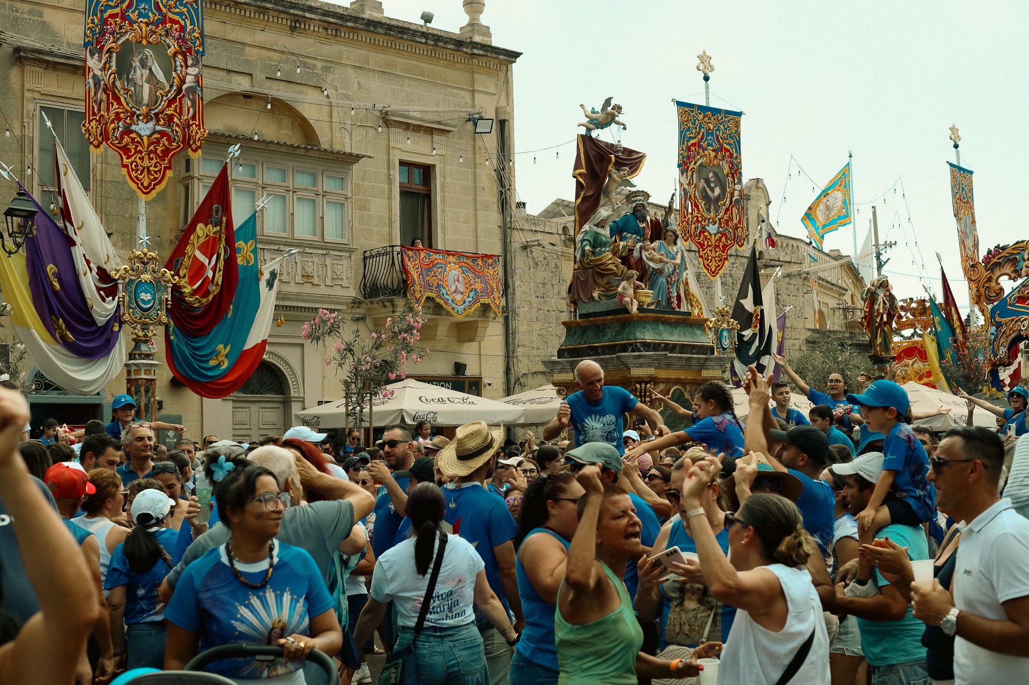 Crowd of people celebrating in a street festival with colorful banners, flags, and religious statues.