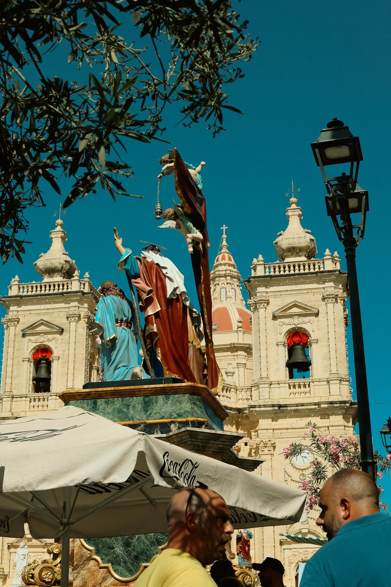 Colorful religious statues and a church with two towers and a dome, with people sitting under an umbrella and a streetlamp nearby