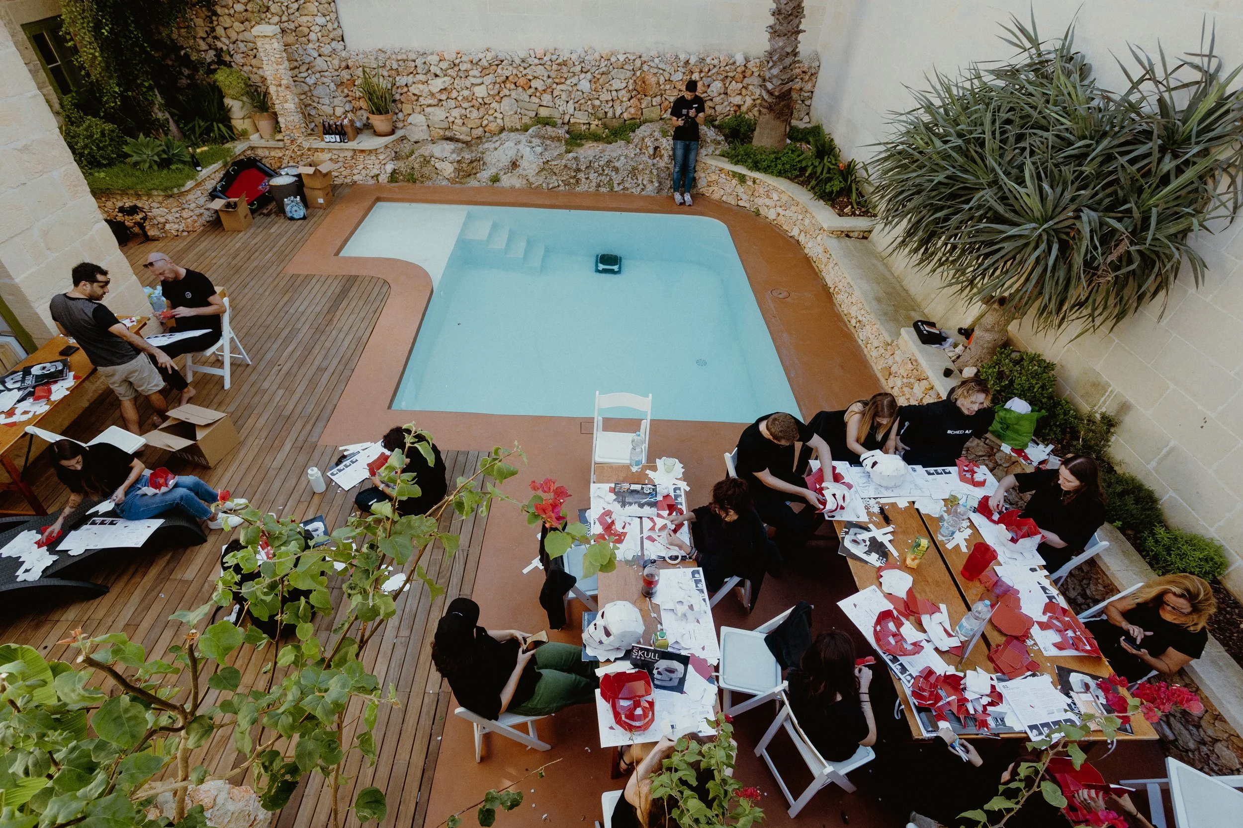 Art Retreat in Malta. A group of people working and socializing around tables near a swimming pool, with some also sitting on a deck and others standing, during the daytime in a backyard with stone walls, plants, and a large tree.