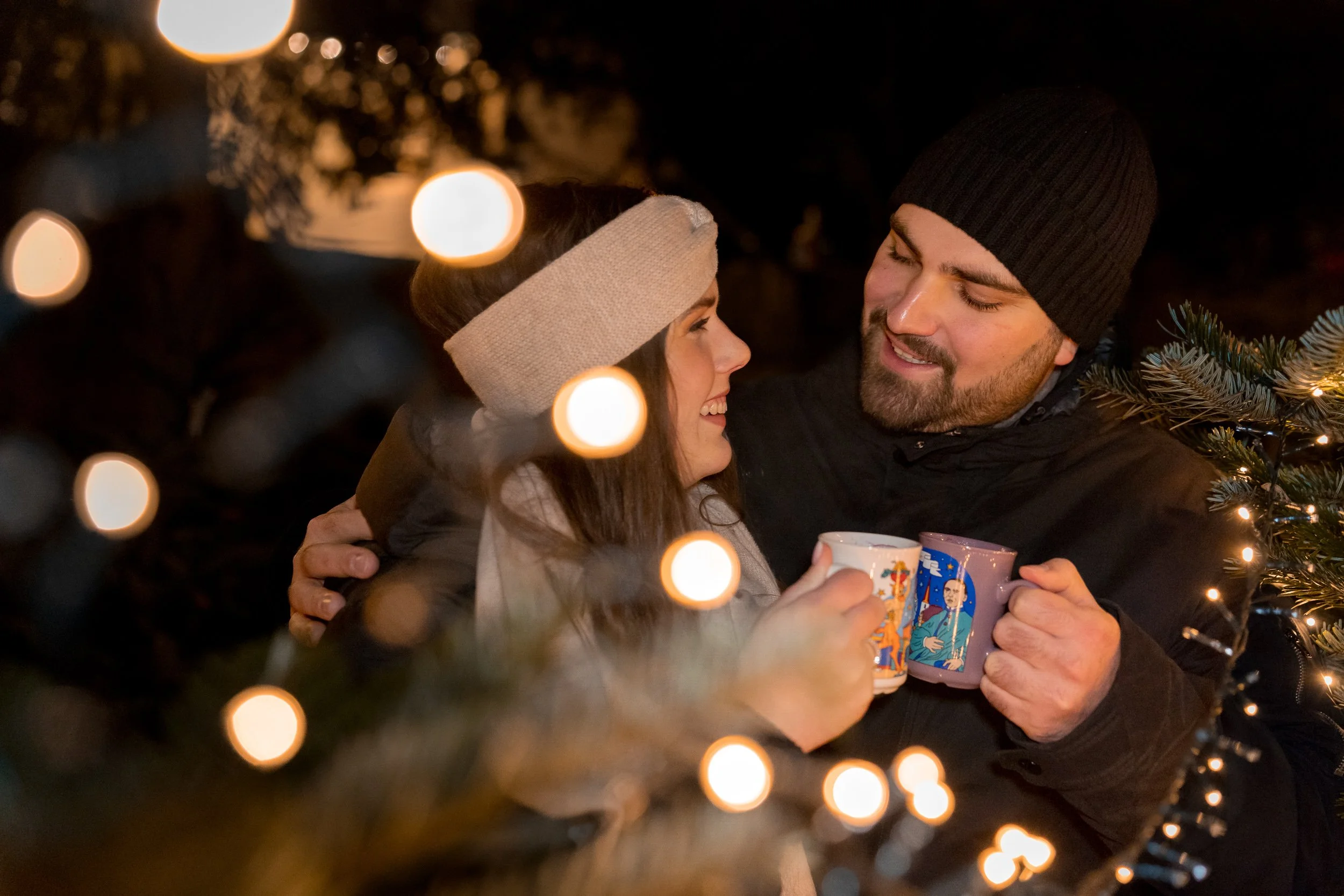 Ein glückliches Paar stößt in winterlicher Umgebung auf dem Weihnachtsmarkt in Deidesheim mit Glühwein-Tassen an, umgeben von Weihnachtslichtern und Tannenzweigen.