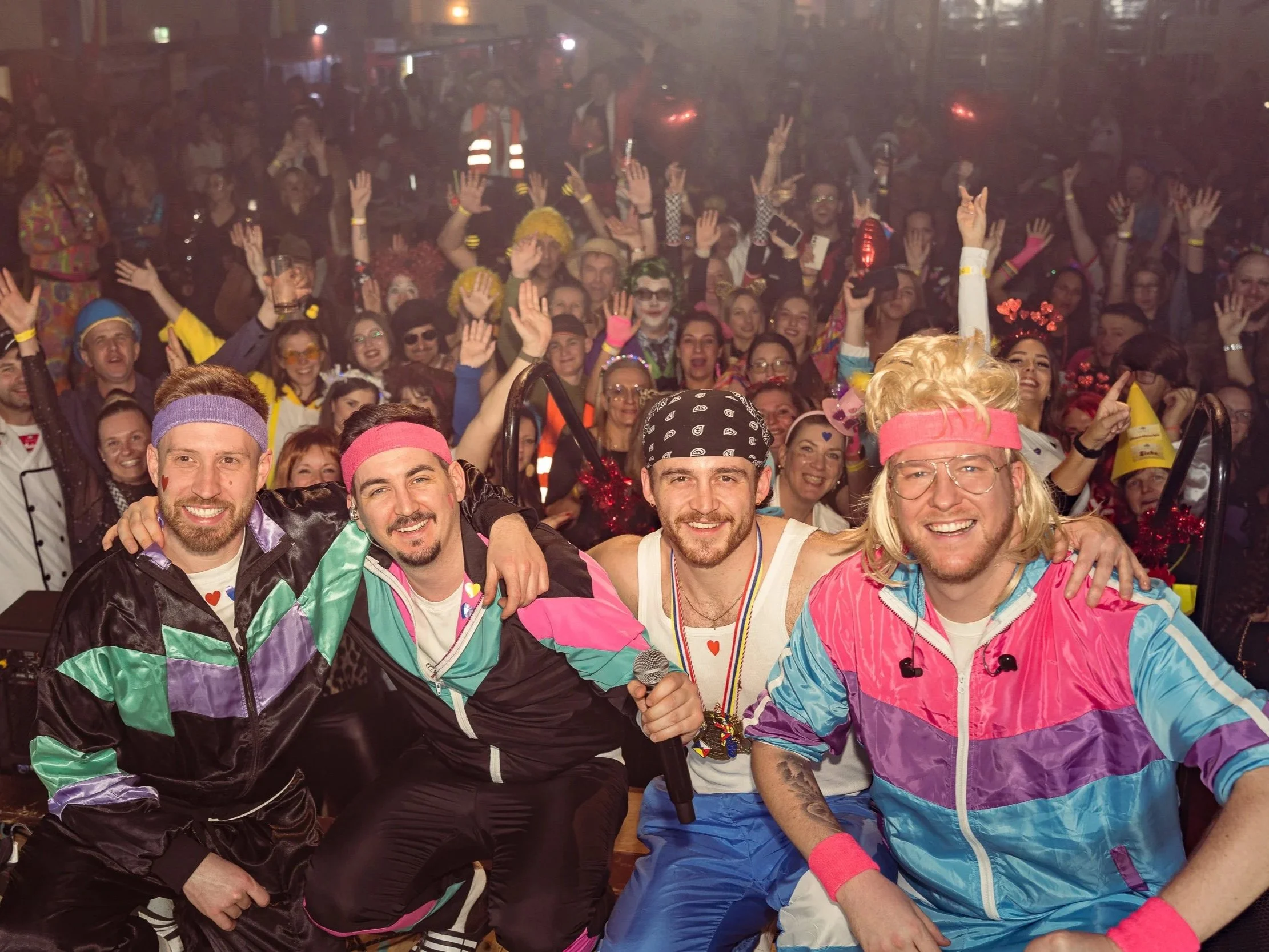 Vier Männer einer Band in bunten 80er-Jahre-Outfits posieren auf einer Bühne in der Festhalle in Oggersheim, umgeben von Menschen in bunten Kostümen bei einer Fasnachts-Party oder Veranstaltung.