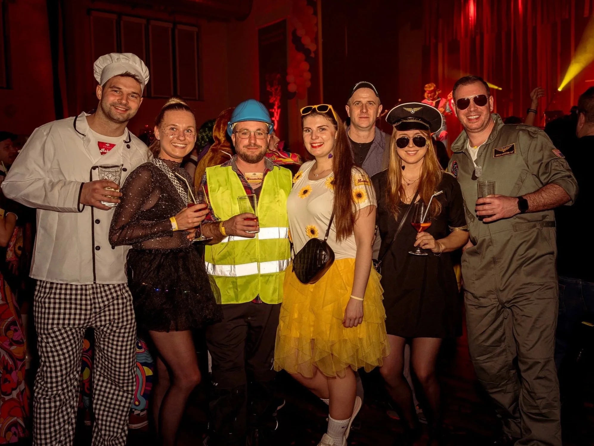 Gruppe von Menschen in Kostümen auf einer Fasnachts-Party, lachend und mit Getränken, im Hintergrund bunte Lichter und Musikbühne.