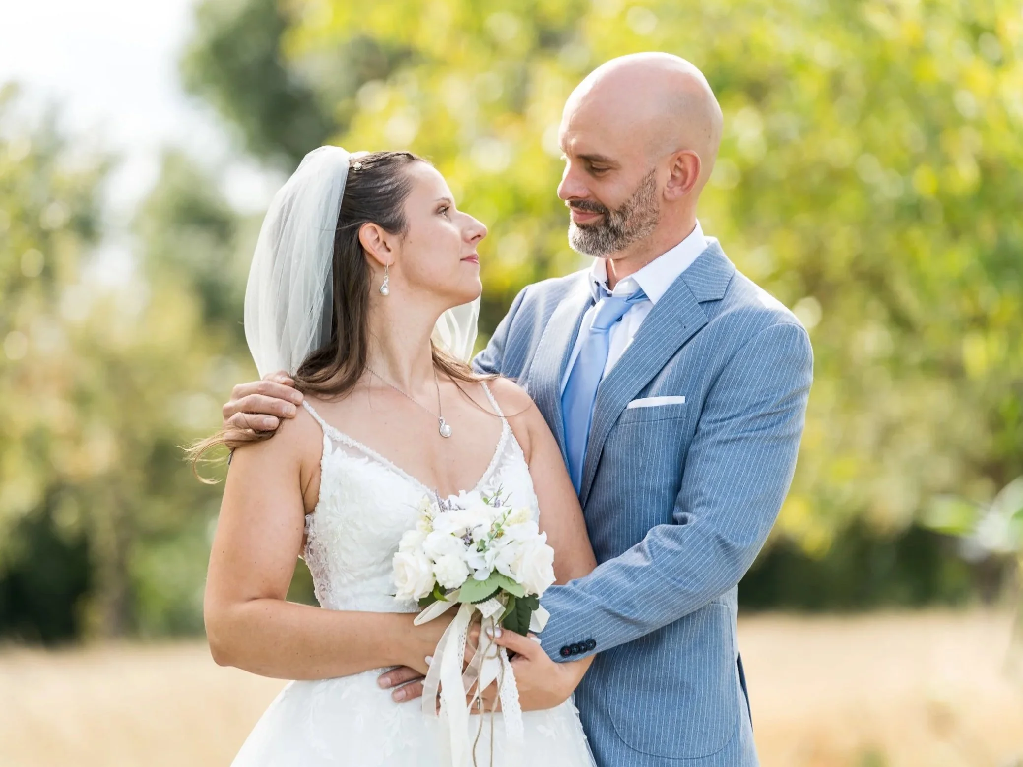 Ein Brautpaar bei einer Hochzeit im Freien in der Pfalz, die Braut trägt ein weißes Kleid und hält einen Blumenstrauß, der Bräutigam trägt einen blauen Anzug, sie schauen sich verliebt an.