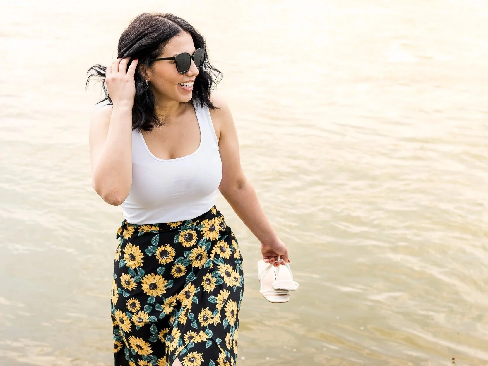 Eine lachende Frau mit dunklen Haaren, Sonnenbrille, weißem Tank-Top, buntem Rock mit Sonnenblumenmuster, steht am Wasser und hält ein Paar weiße Schuhe in der Hand.