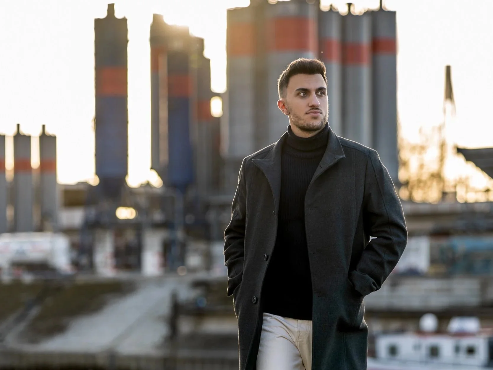 Junger Mann mit kurzen schwarzen Haaren trägt schwarzen Mantel Jacke und helle Hose, steht im Mannheimer Hafengelände vor einer Industrielandschaft bei Sonnenuntergang.