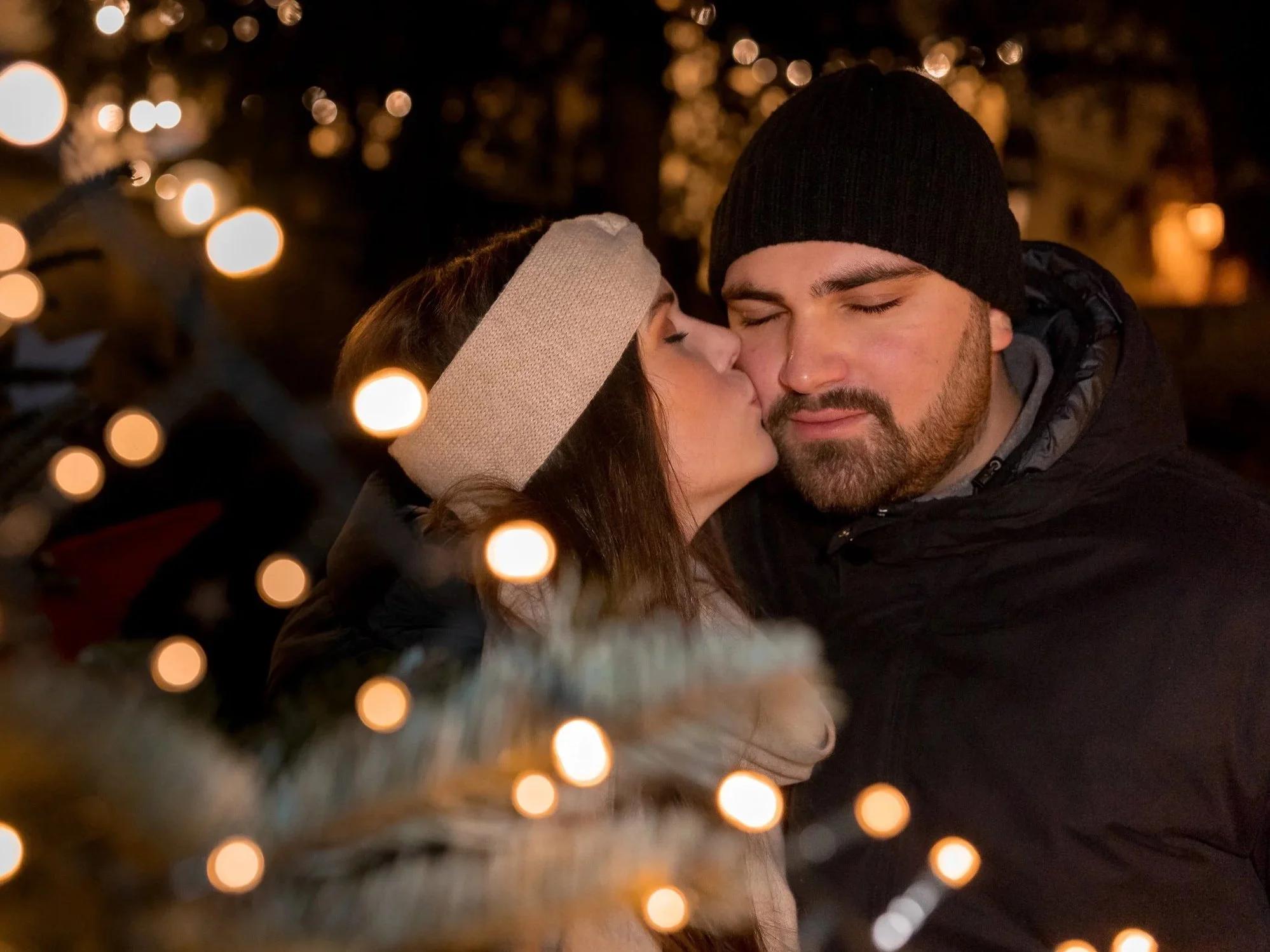 Ein glückliches Paar küsst sich in winterlicher Umgebung auf dem Weihnachtsmarkt in Deidesheim, umgeben von Weihnachtslichtern und Tannenzweigen.