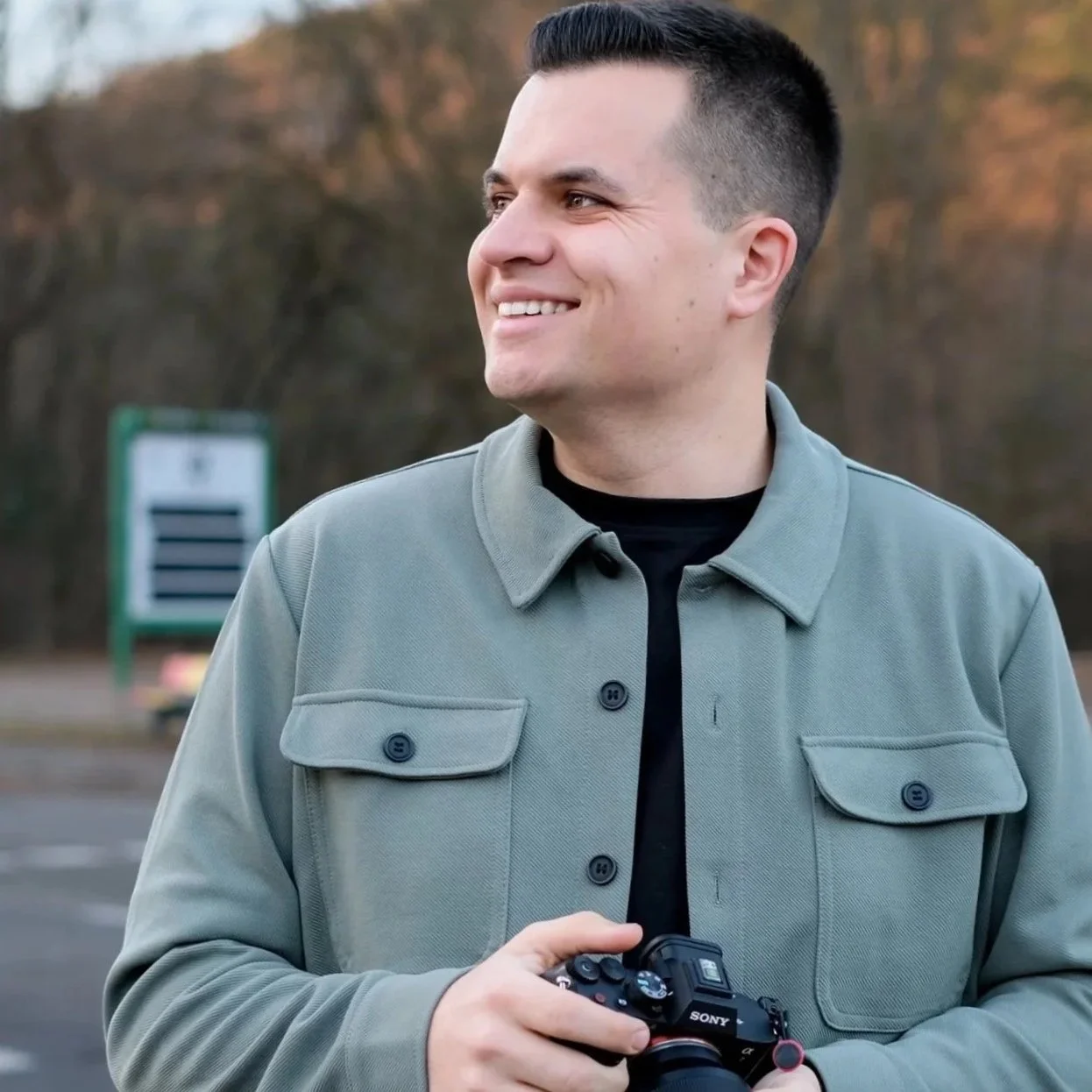 Ein Mann mit kurzem schwarzen Haar, lächelnd, trägt eine graue Jacke und hält eine Sony Kamera in der Hand, steht im Freien vor einem unscharfen Hintergrund mit Bäumen und einem grünen Schild.
