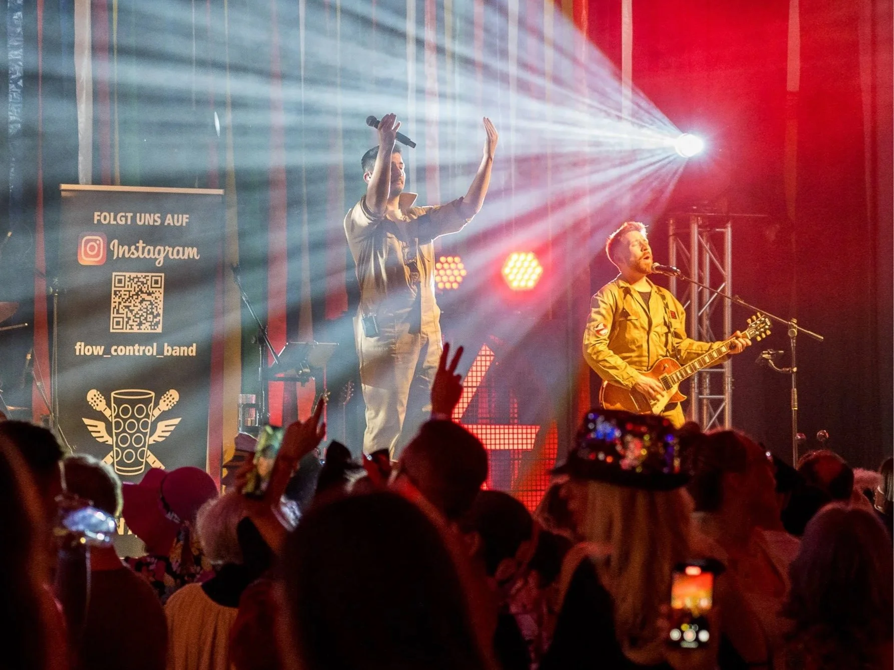 Zwei Musiker auf einer Bühne mit rotem Beleuchtung, einer singt und spielt Gitarre, der andere hält ein Mikrofon und hebt die Hand, im Publikum sind Menschen und eine Person trägt eine Party- oder Aufführungsmütze.