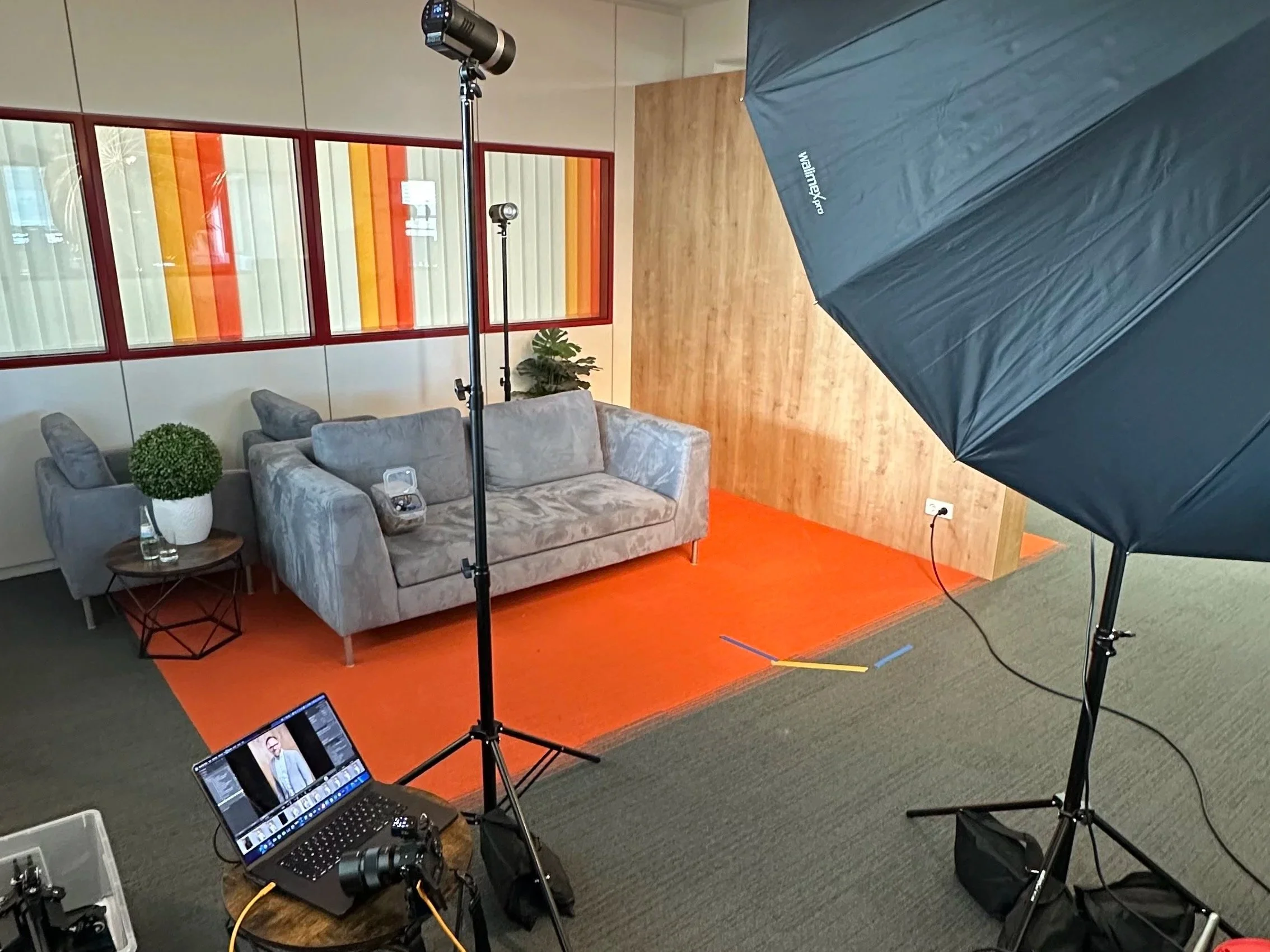 Shooting-Setup mit grauem Sofa, Pflanzen, Fotoausrüstung, Lichtreflektor, Laptop, Kamera und Hintergrundvorbereitungen für Video- oder Fotoshooting in einem Raum.