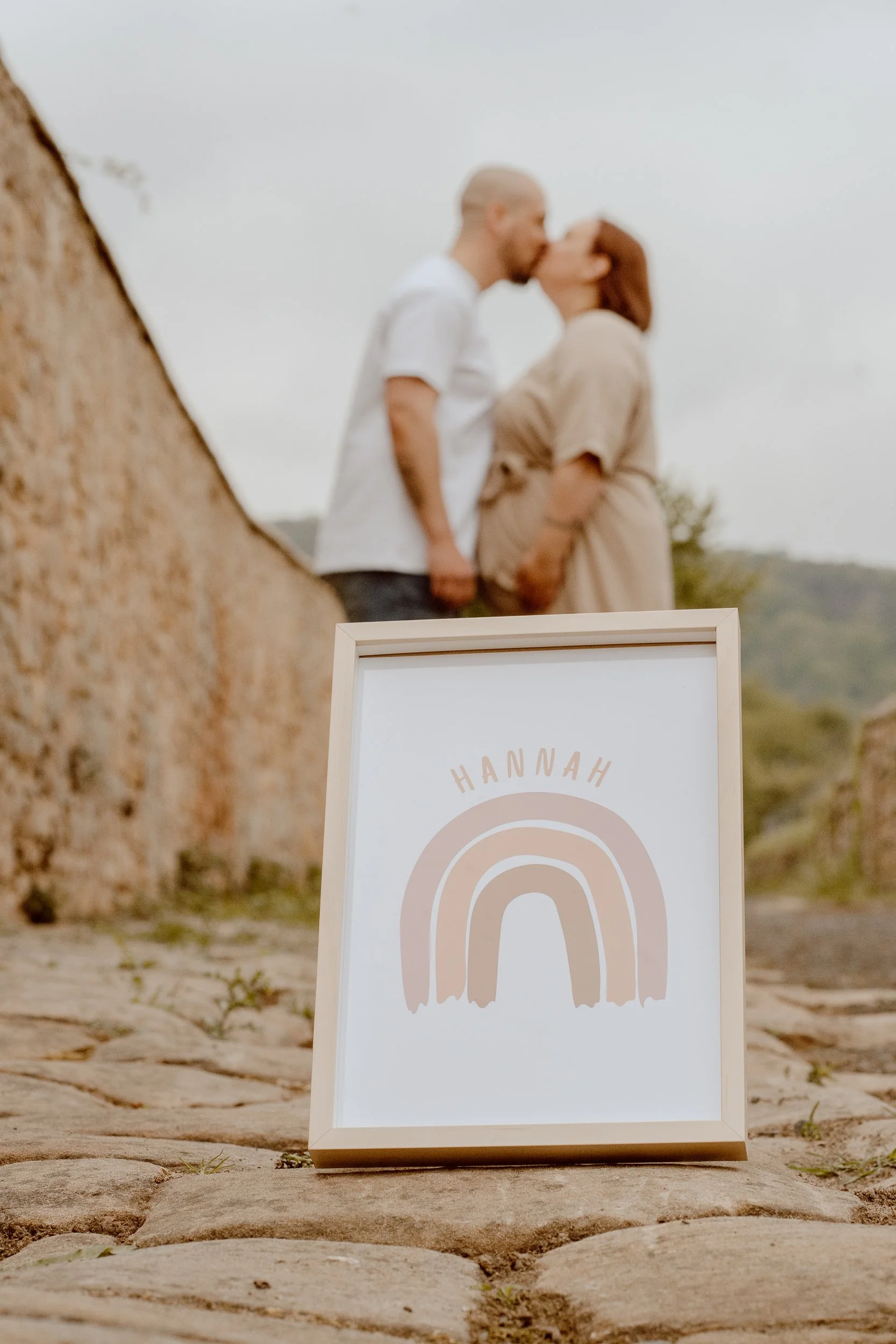 Ein Holzkarton mit dem Namen 'Hannah' und einem Regenbogen-Design, im Vordergrund. Im Hintergrund ein Paar, das sich küsst, auf einer Steinmauer an der Weinstraße im Freien.