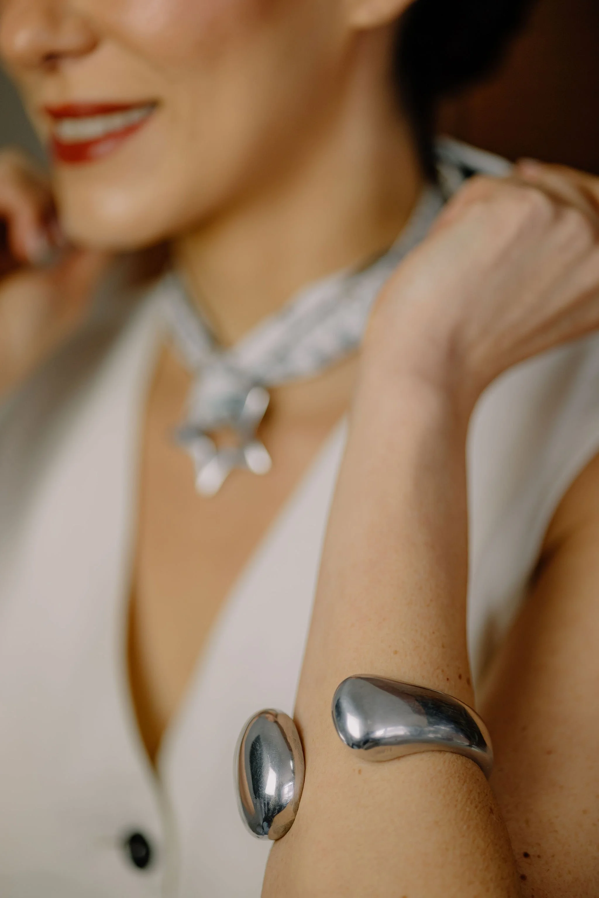 Close-up of a woman smiling, showing her red lipstick, wearing a necklace and a white sleeveless top with black buttons, and a metallic bracelet on her arm.