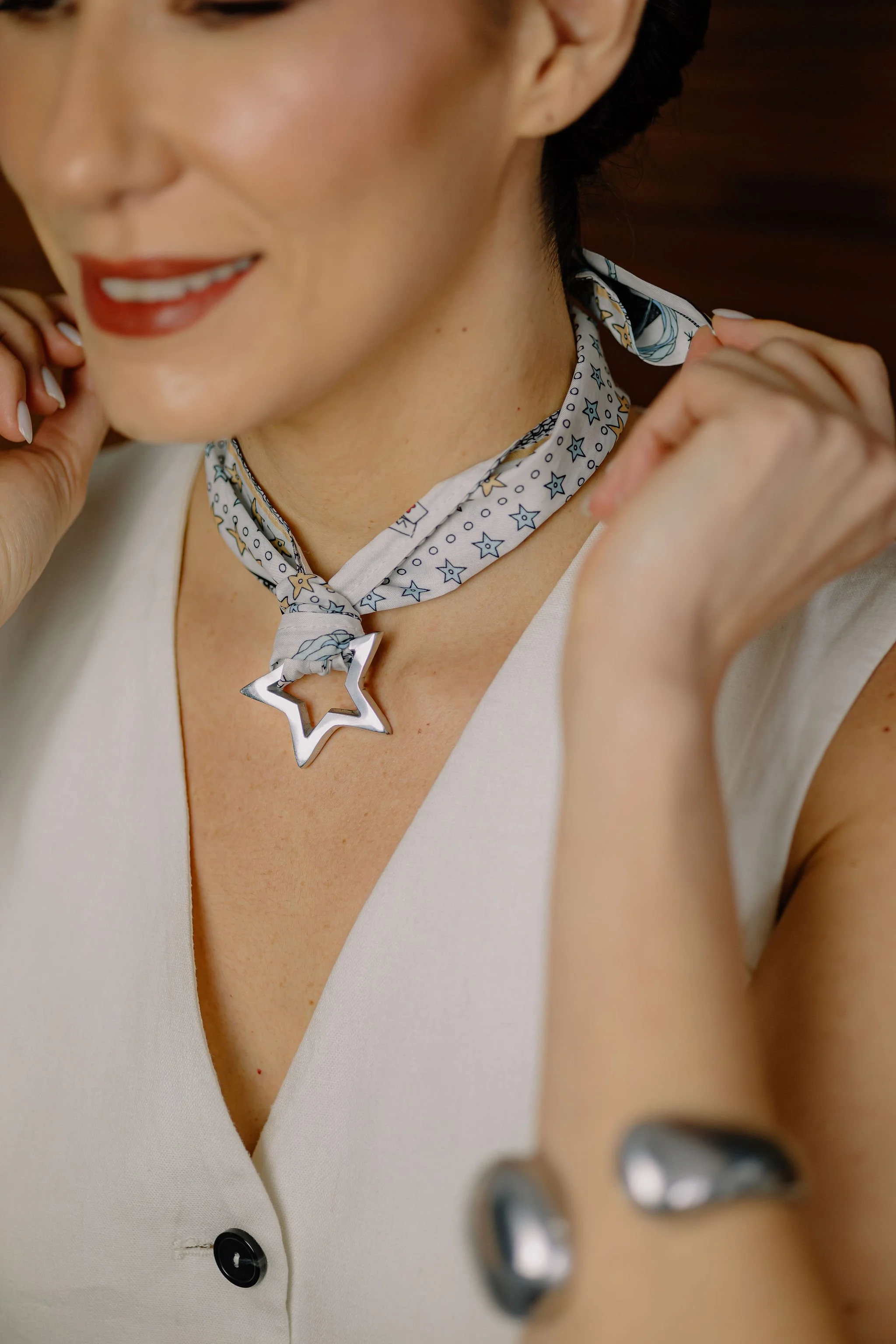 Close-up of a woman adjusting a star-shaped necklace, wearing a patterned scarf, sleeveless white top, and smiling with red lipstick.