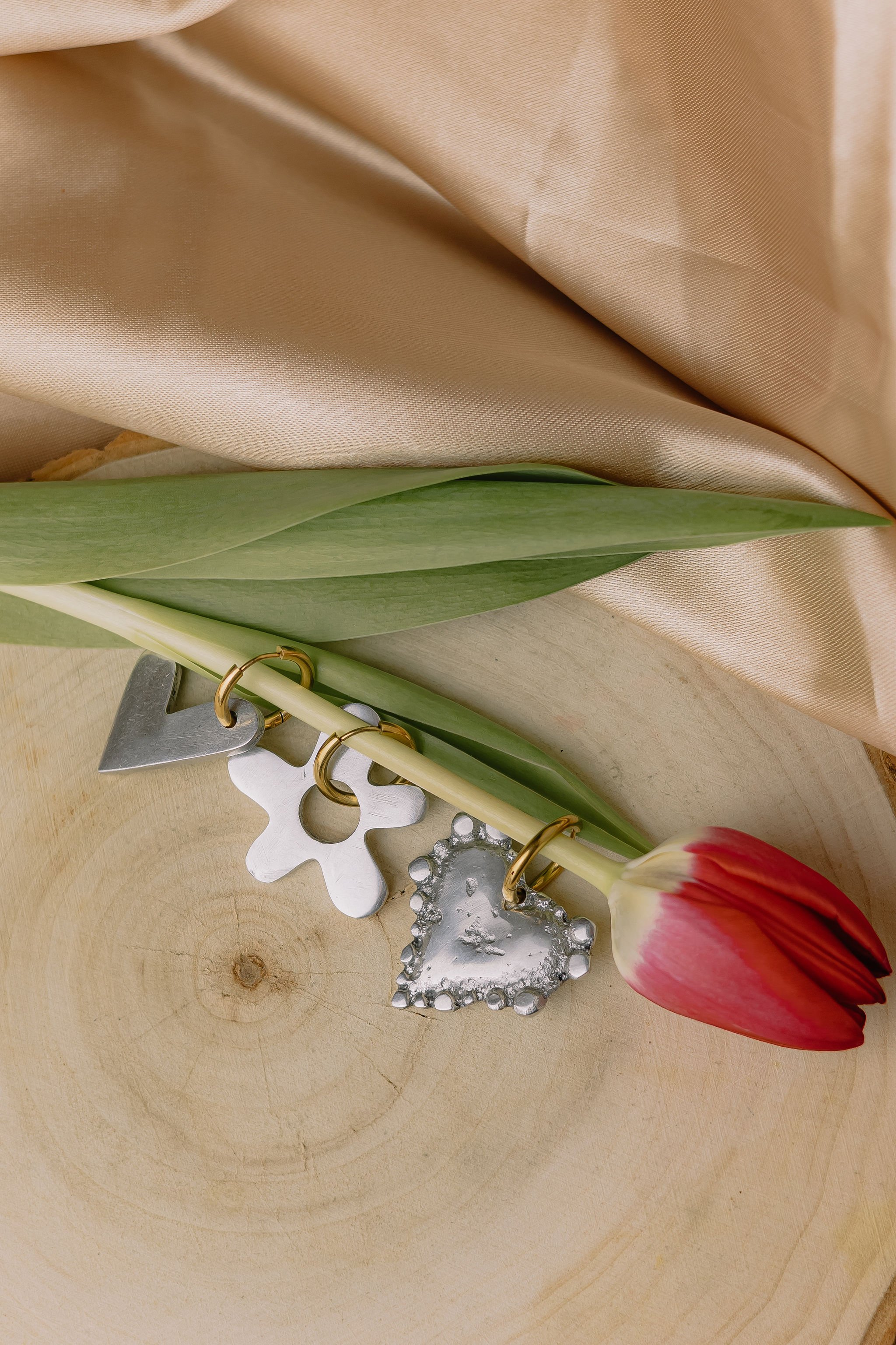A pink and white tulip flower with green leaves, a piece of beige satin fabric, and decorative metal heart and gear charms placed on a wooden surface.
