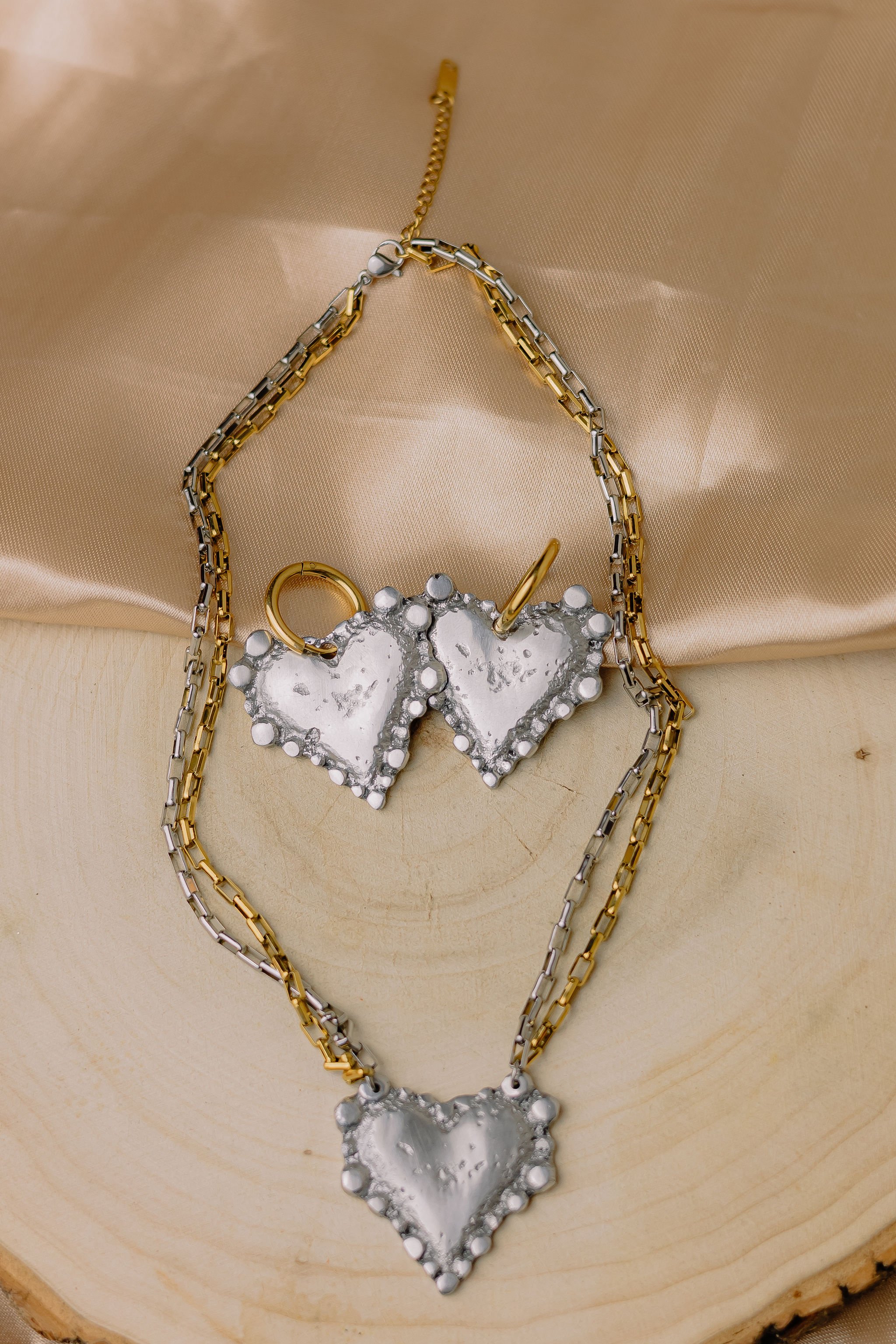 Silver heart-shaped pendant with a beaded border on a silver and gold chain, placed on a wooden surface with gold satin fabric in the background.