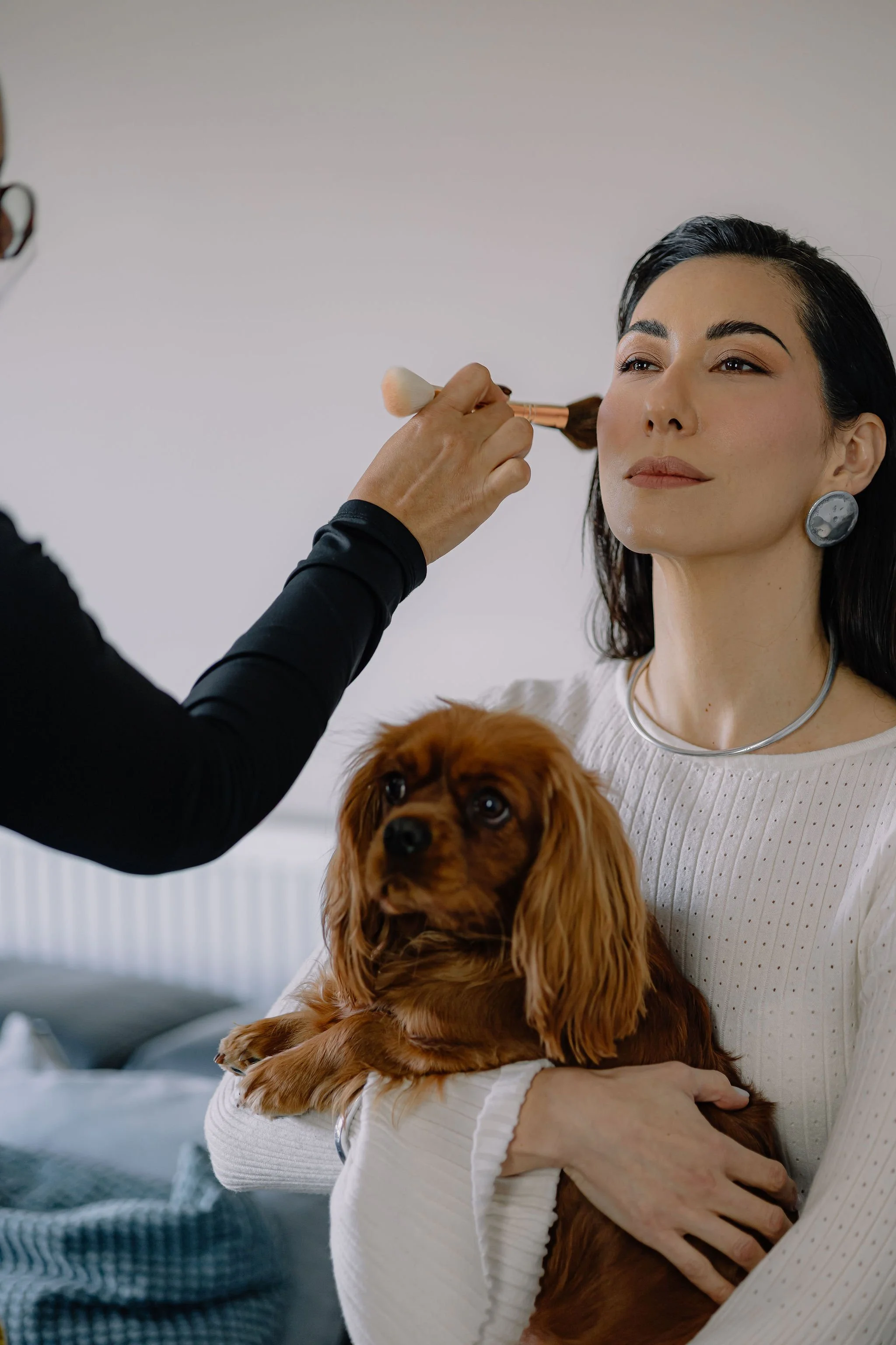 A woman with dark hair and earrings is holding a small brown dog while another person applies makeup with a brush to her face.