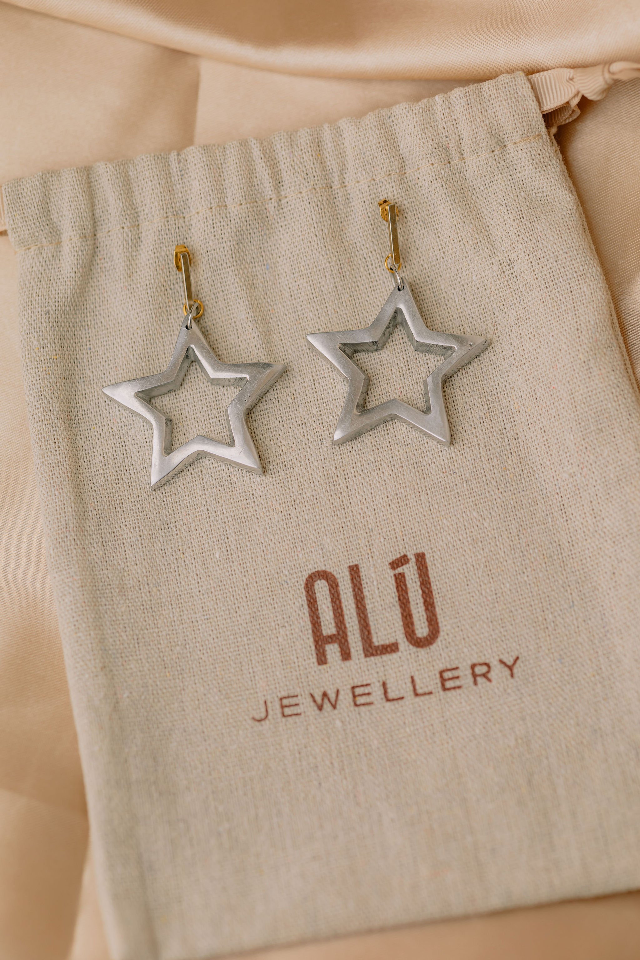 Silver star-shaped earrings hanging on a beige fabric pouch with the brand name 'ALÜ Jewellery' printed on it.