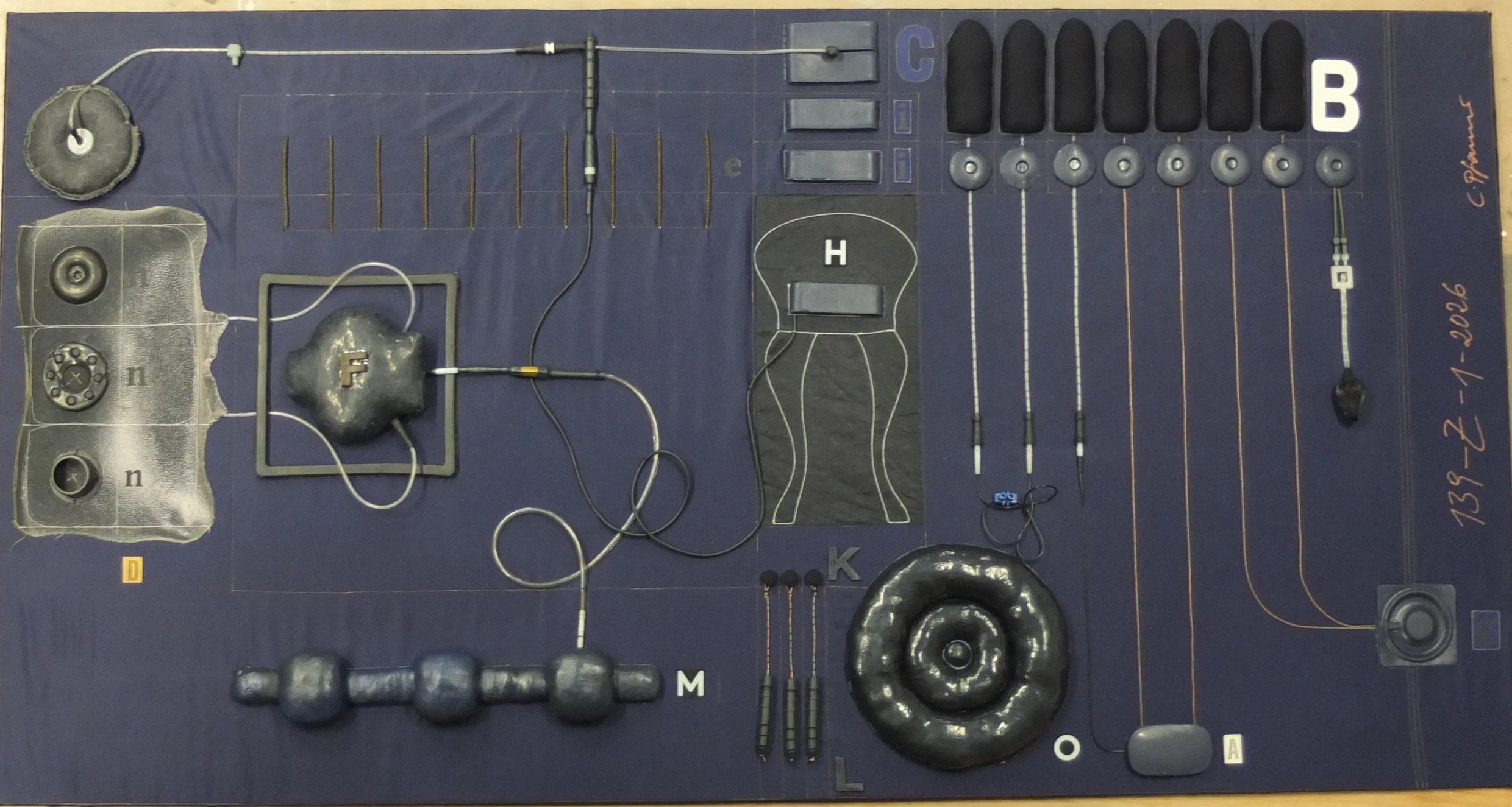 Ein graphisches Kunstwerk mit verschiedenen metallischen und textilen Elementen, verbunden durch Drähte und Kabel, mit Buchstaben, Nummern und Linien, die ein komplexes Layout bilden.