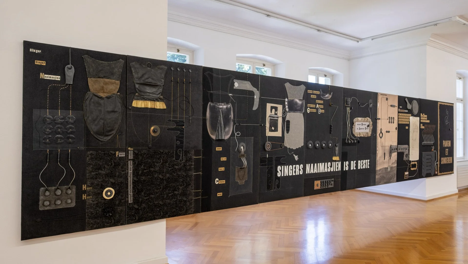 Eine Wand mit einer künstlerischen Installation, die verschiedene Gegenstände und Materialien in Schwarz, Grau und Beige enthält, mit Text in Niederländisch und Deutsch, der auf Sängern und Nägeln Bezug nimmt.
