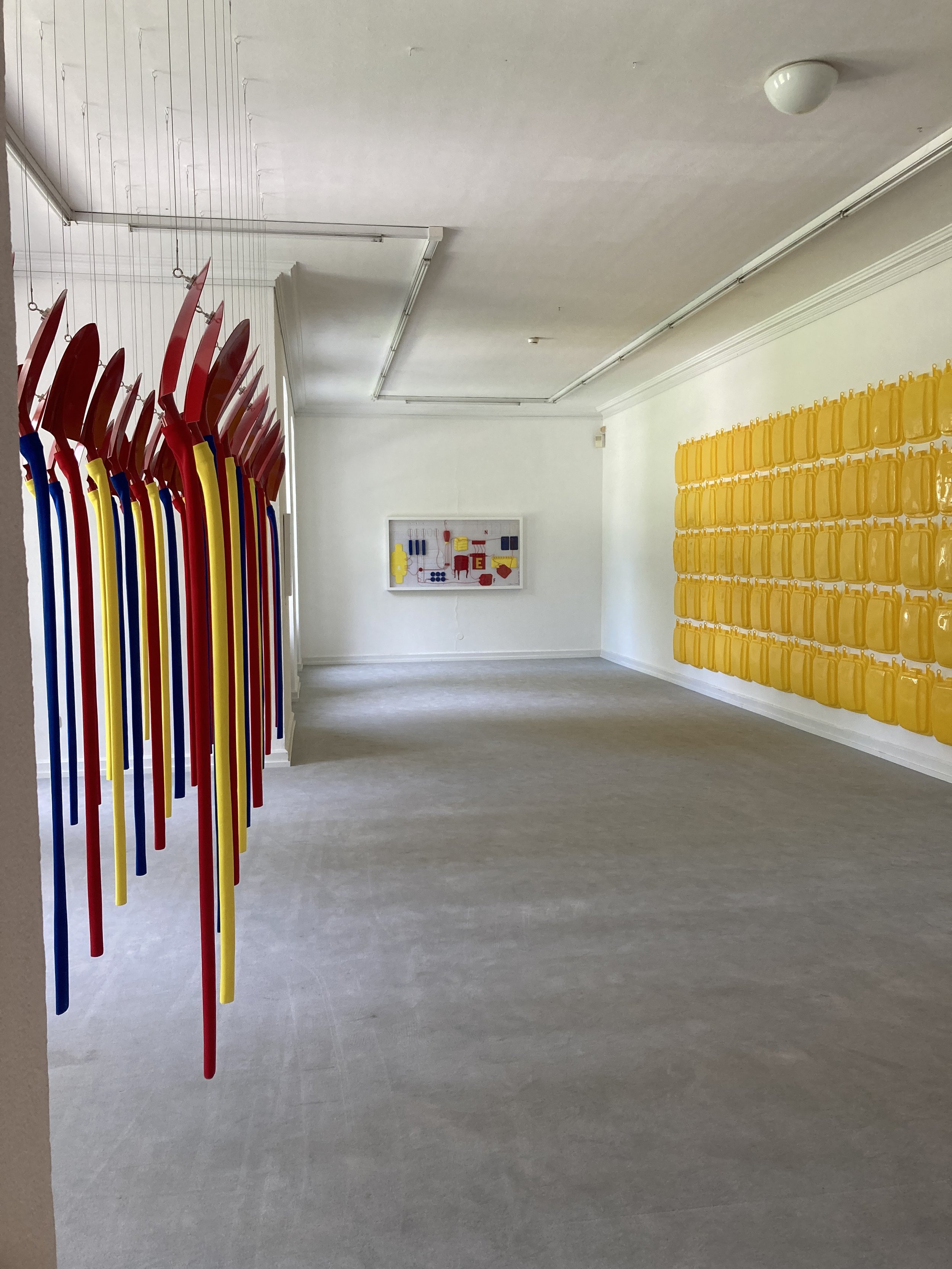 Ein Raum mit mehreren in Rot, Blau, Gelb und Schwarz gestrichenen Stuhllen, einer weißen Wand mit einer Kunstinstallation und einer Wand mit gelben Hängeregalen.