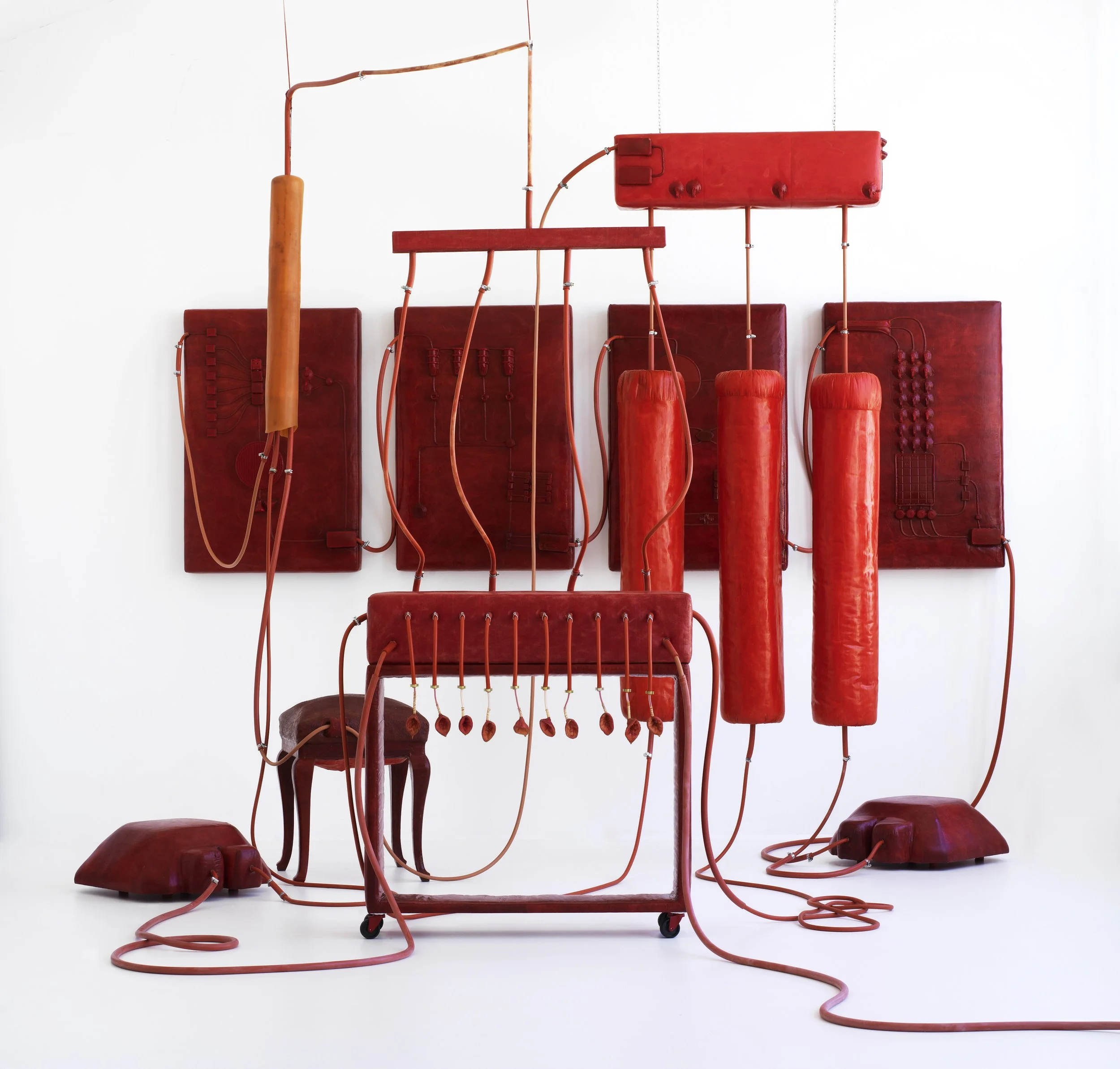 Kunstinstallation aus roten elektronischen Komponenten, Kabeln, Kupfern, auf einer weißen Wand, kombiniert mit einem alten Holstuhl als Teil des Kunstwerks.