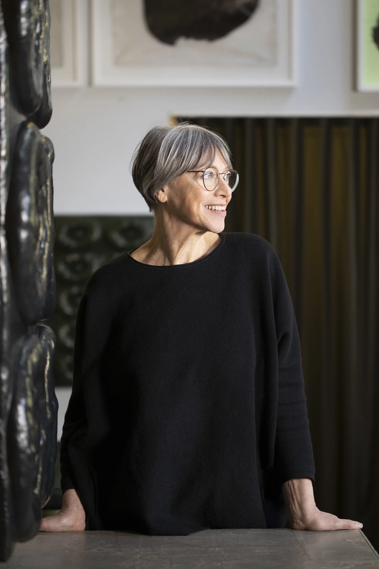 Eine ältere Frau mit kurzen grauen Haaren, runder Brille und schwarzem Pullover steht hinter einem Tisch, ihre Hände ruhen auf der Oberfläche. Sie lächelt während sie nach rechts schaut, im Hintergrund sind Kunstwerke auf der Wand sichtbar.