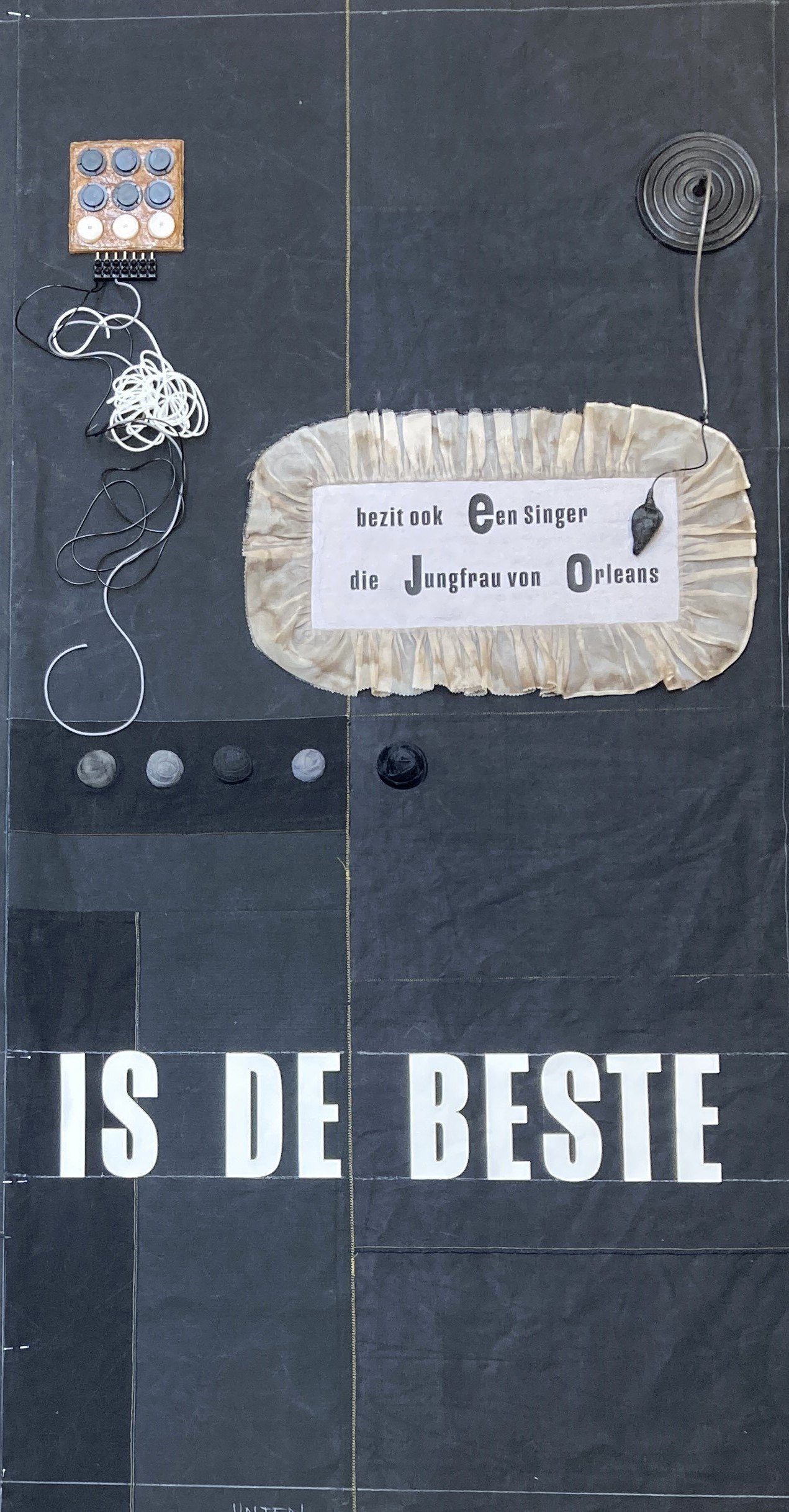 Ein Wandbild mit einem weißen Stoffbanner, auf dem geschrieben steht: "bezir ook een Singer die Jungfrau von Orleans" und darunter große weiße Buchstaben: "IST DE BESTE" auf einem dunklen Hintergrund mit verschiedenen Gegenständen wie Knöpfen, Kabeln und einem runden Gegenstand.