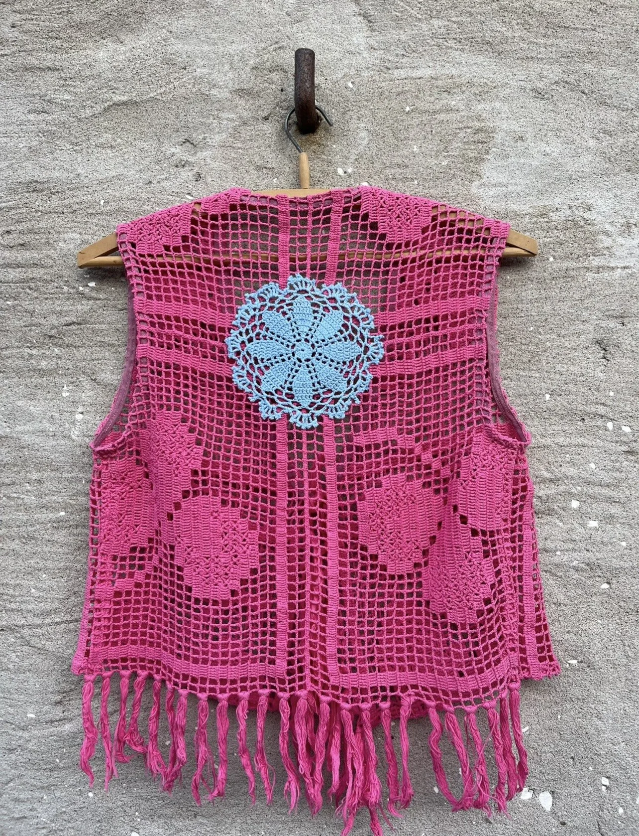 Pink crocheted vest with fringe at the bottom, featuring a large blue crochet flower in the center, hanging on a wooden hanger against a concrete wall.