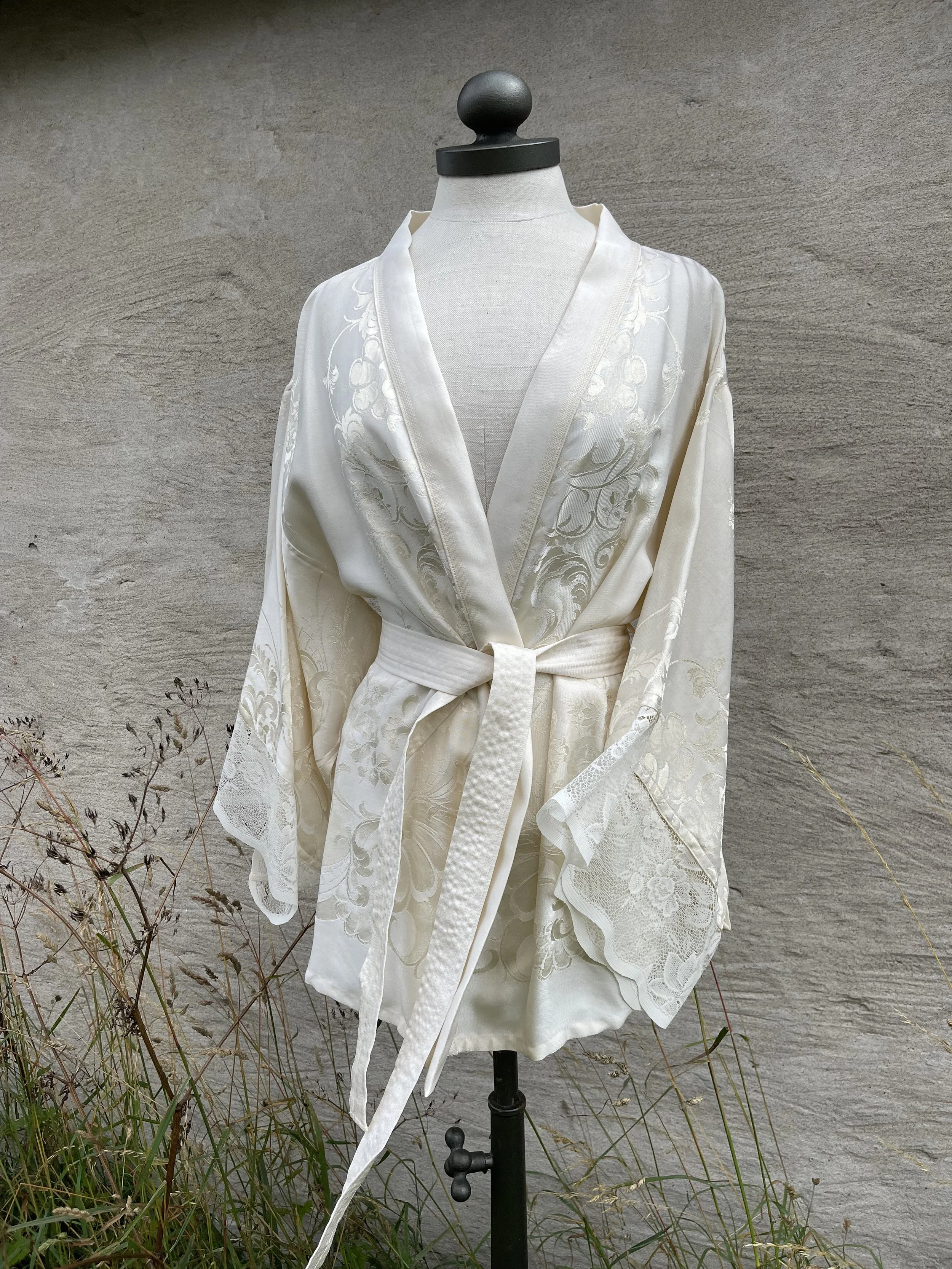 White satin robe with floral embroidery and lace details, tied at the waist, displayed on a dress form in front of a stone wall.