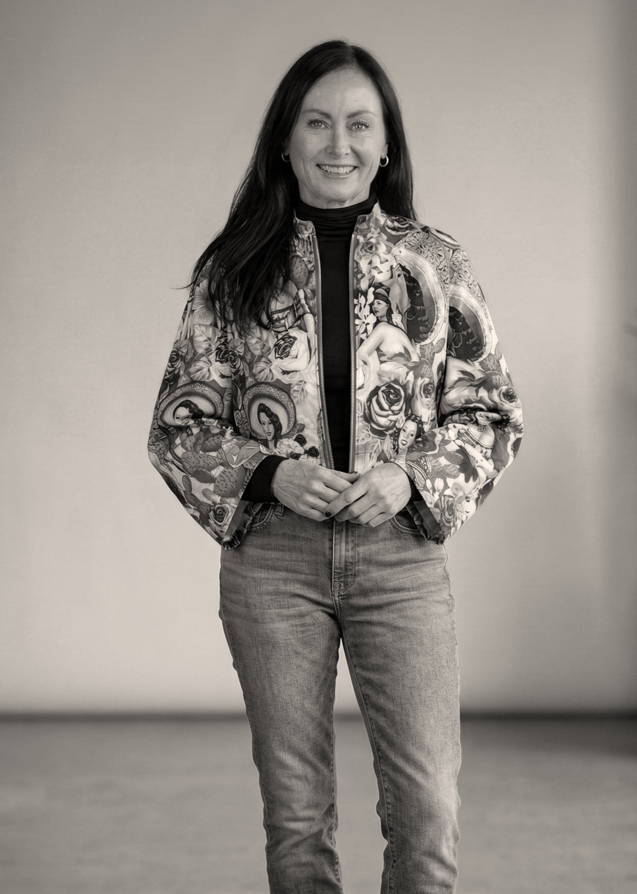 A woman with long dark hair smiling and standing against a plain background, wearing a colorful patterned jacket and jeans.