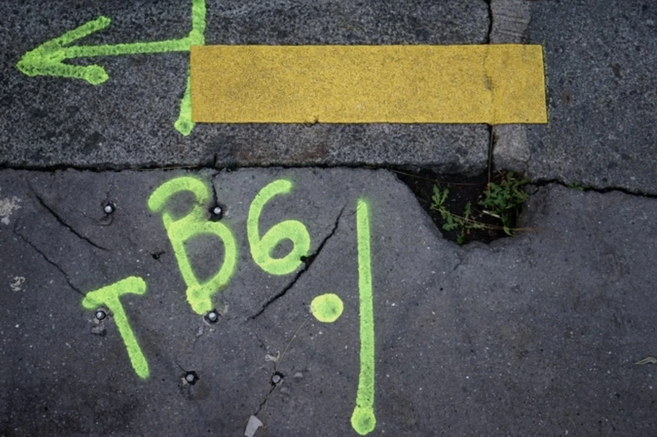 Street pavement with neon green spray-painted text 'T B6.1' and an arrow pointing to a yellow tape strip, with some small green plants growing in a crack.