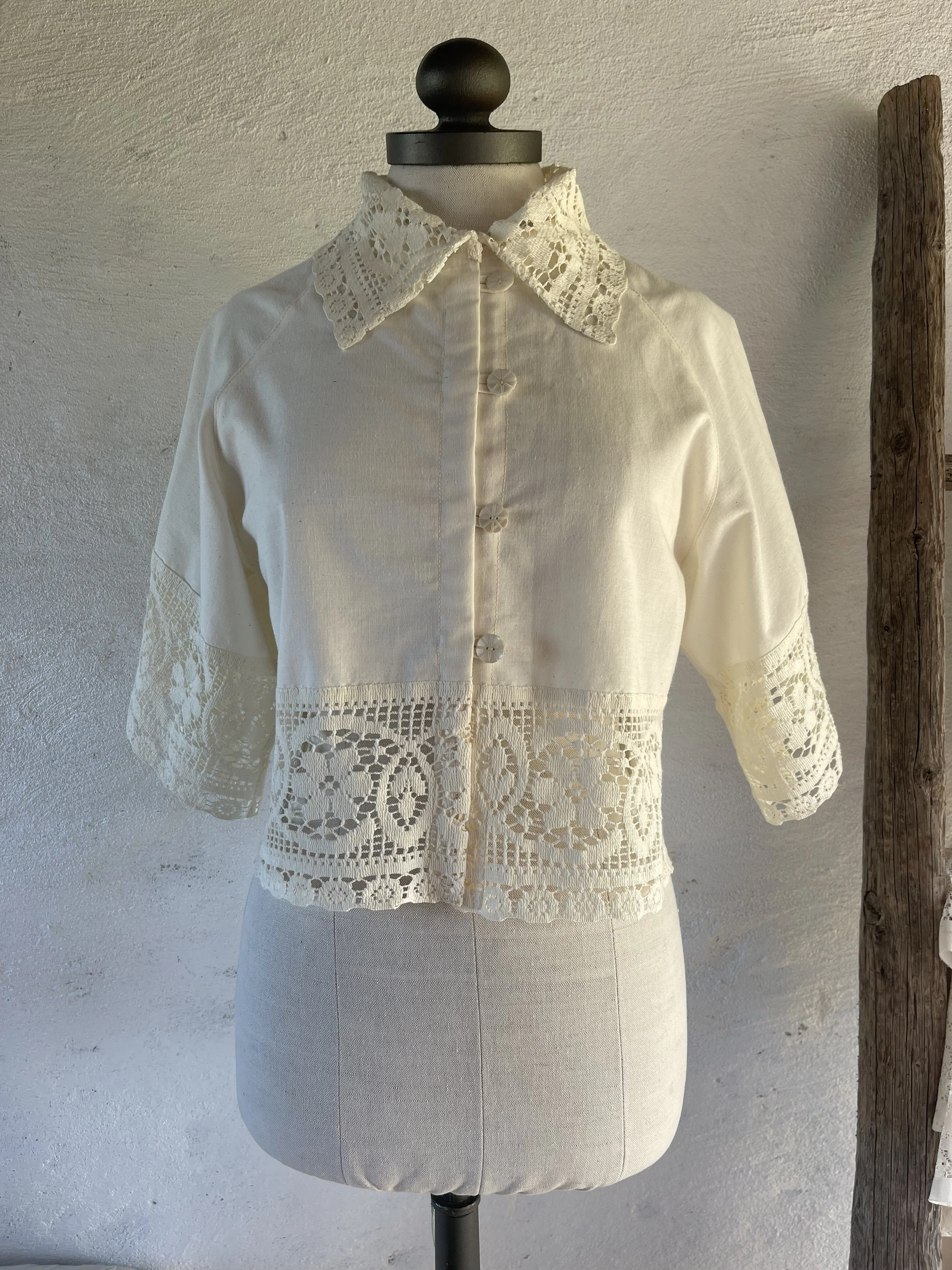 Cream-colored vintage blouse with lace detailing and embroidered patterns, displayed on a mannequin against a plain wall.