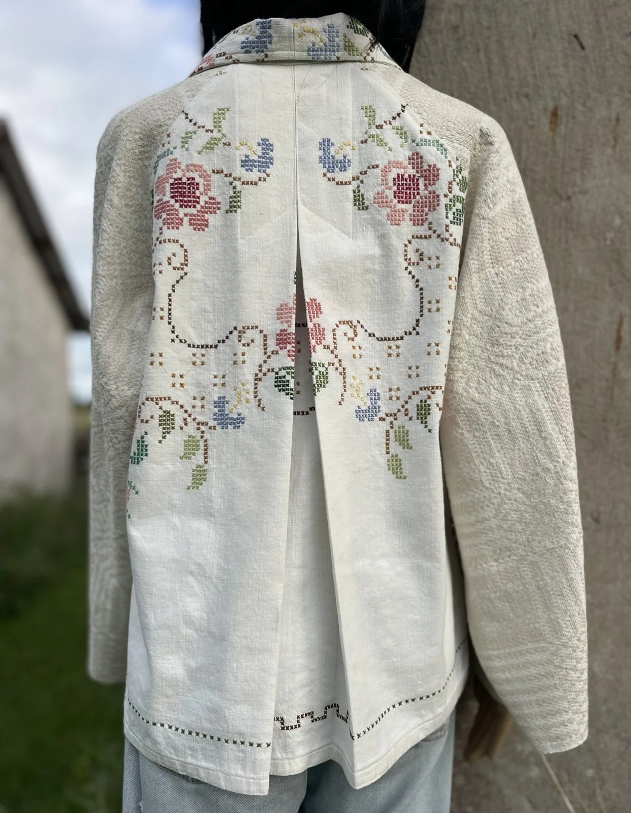 Back of a white embroidered jacket with floral and vine patterns in pink, blue, green, and brown, worn over a shirt and gray pants, outdoors near a stone wall.