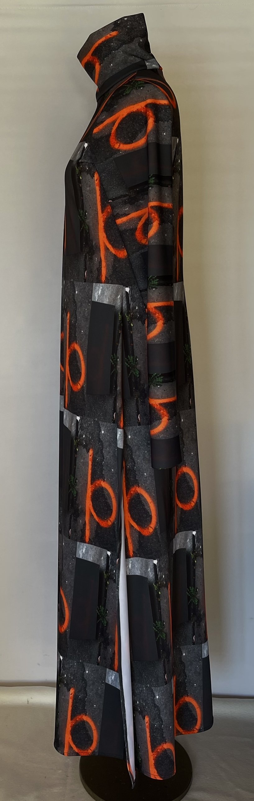 A mannequin dressed in a long coat with a black background and a pattern of neon orange circles and green plants printed all over, standing against a plain white wall.