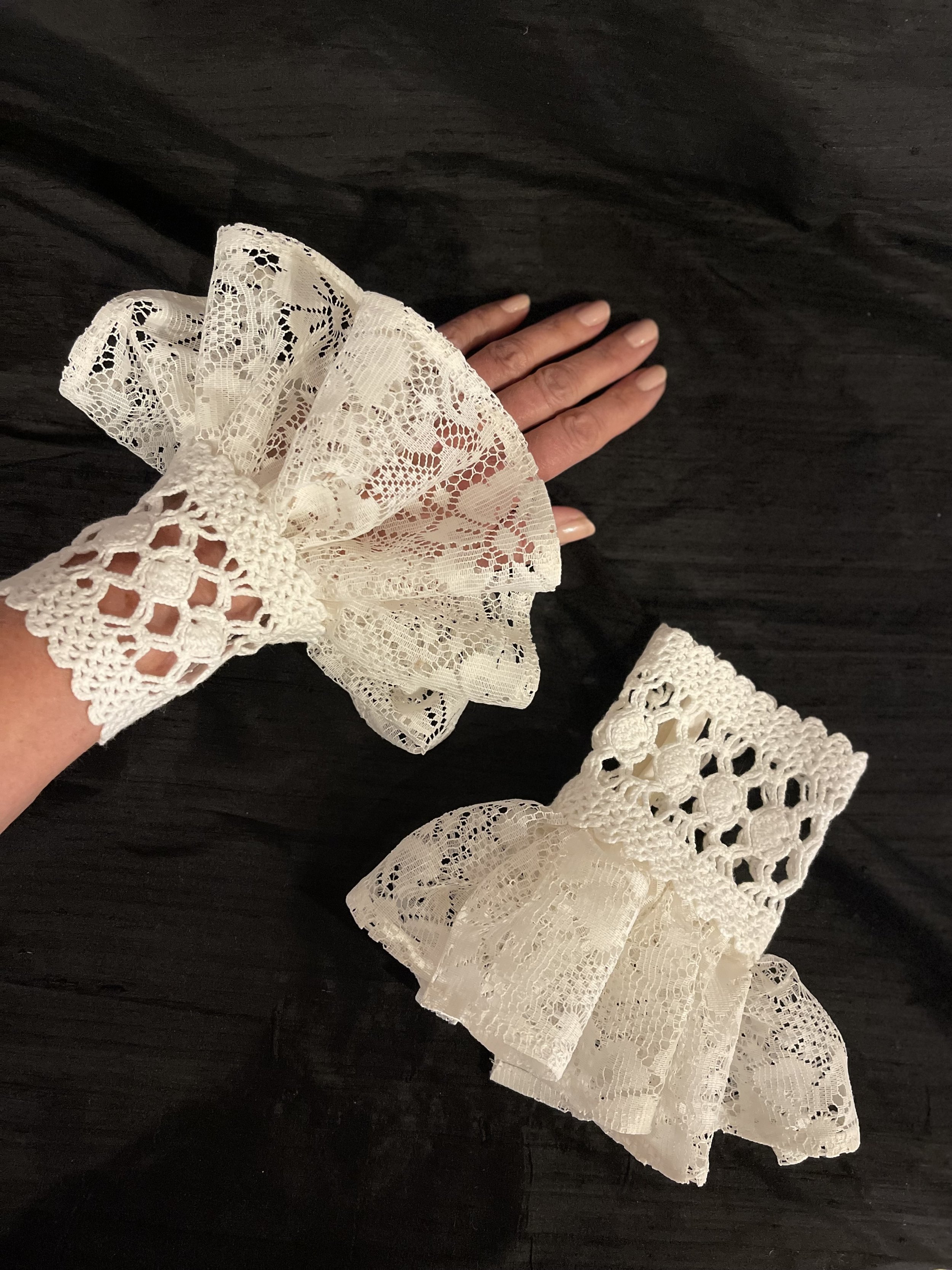 Hand wearing a large white lace cuff with ruffled design, alongside an identical lace cuff lying flat on a dark surface.