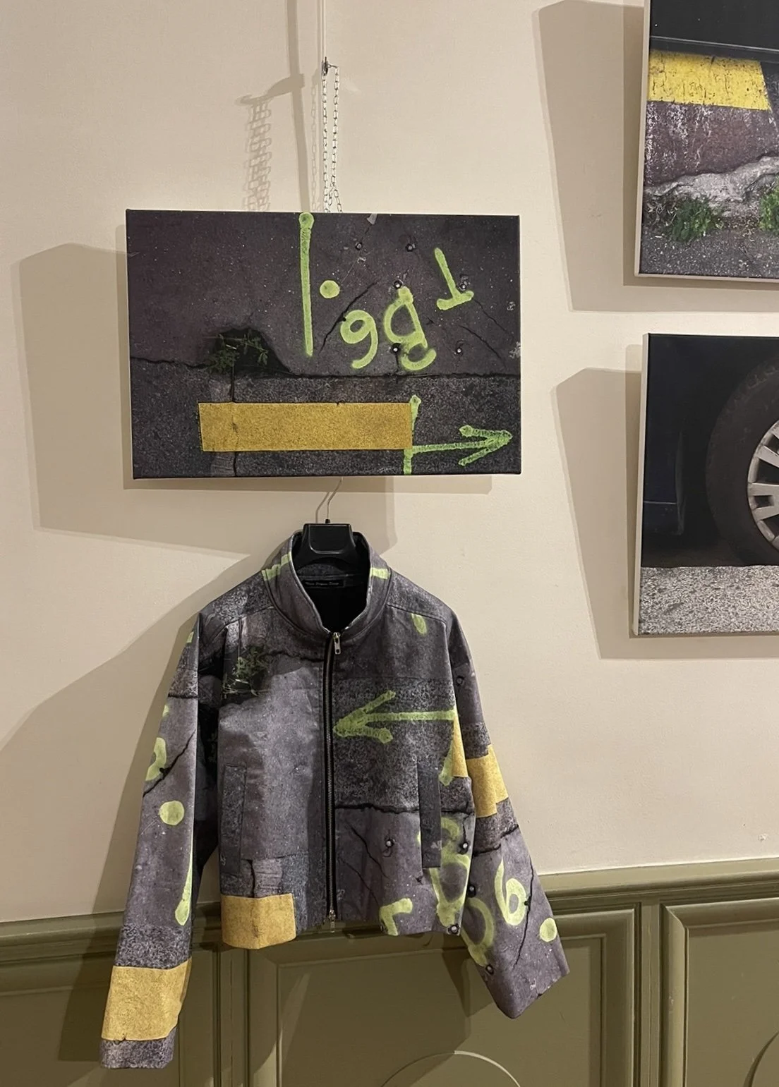 A wall display featuring street art-inspired clothing and artwork. There is a jacket with a concrete wall and spray-painted yellow markings, along with a matching canvas above it showing graffiti and directional arrows.