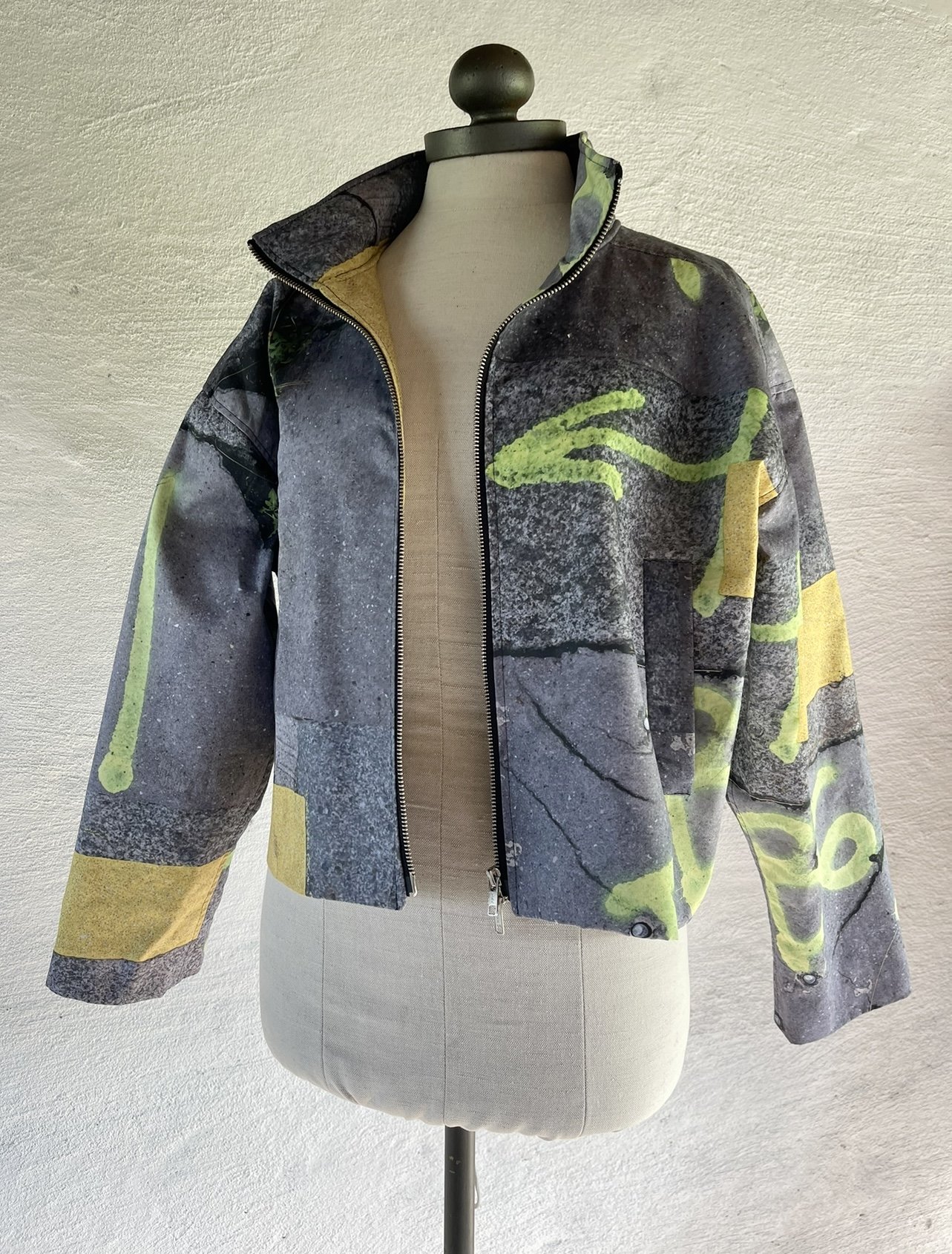 A jacket with a design resembling a concrete wall with yellow and green graffiti, displayed on a mannequin. 