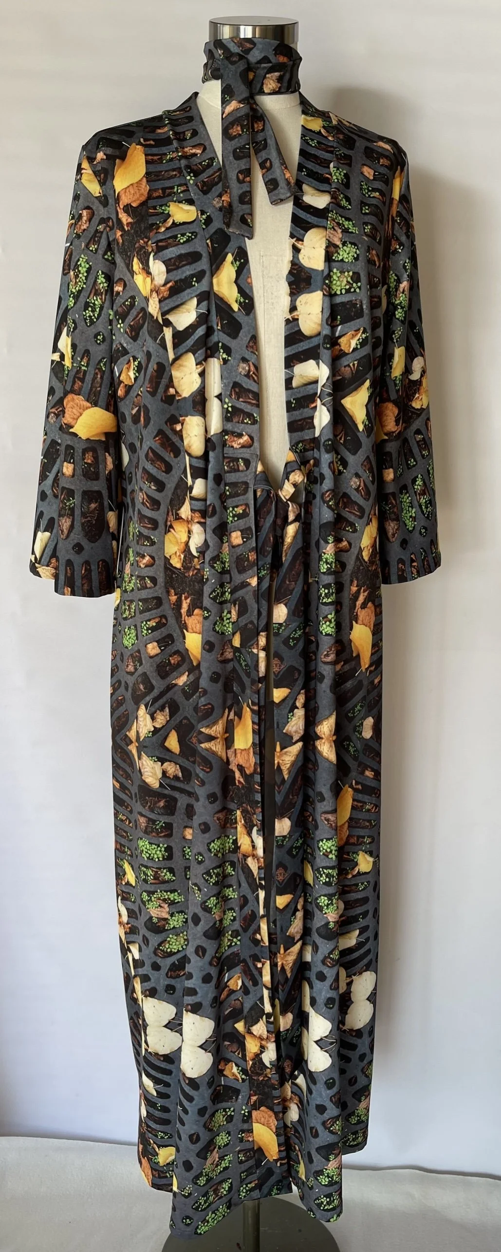 A long, black dress with a cutout pattern resembling gills and featuring a print of various vegetables, including yellow squash, mushrooms, and green herbs, displayed on a mannequin.