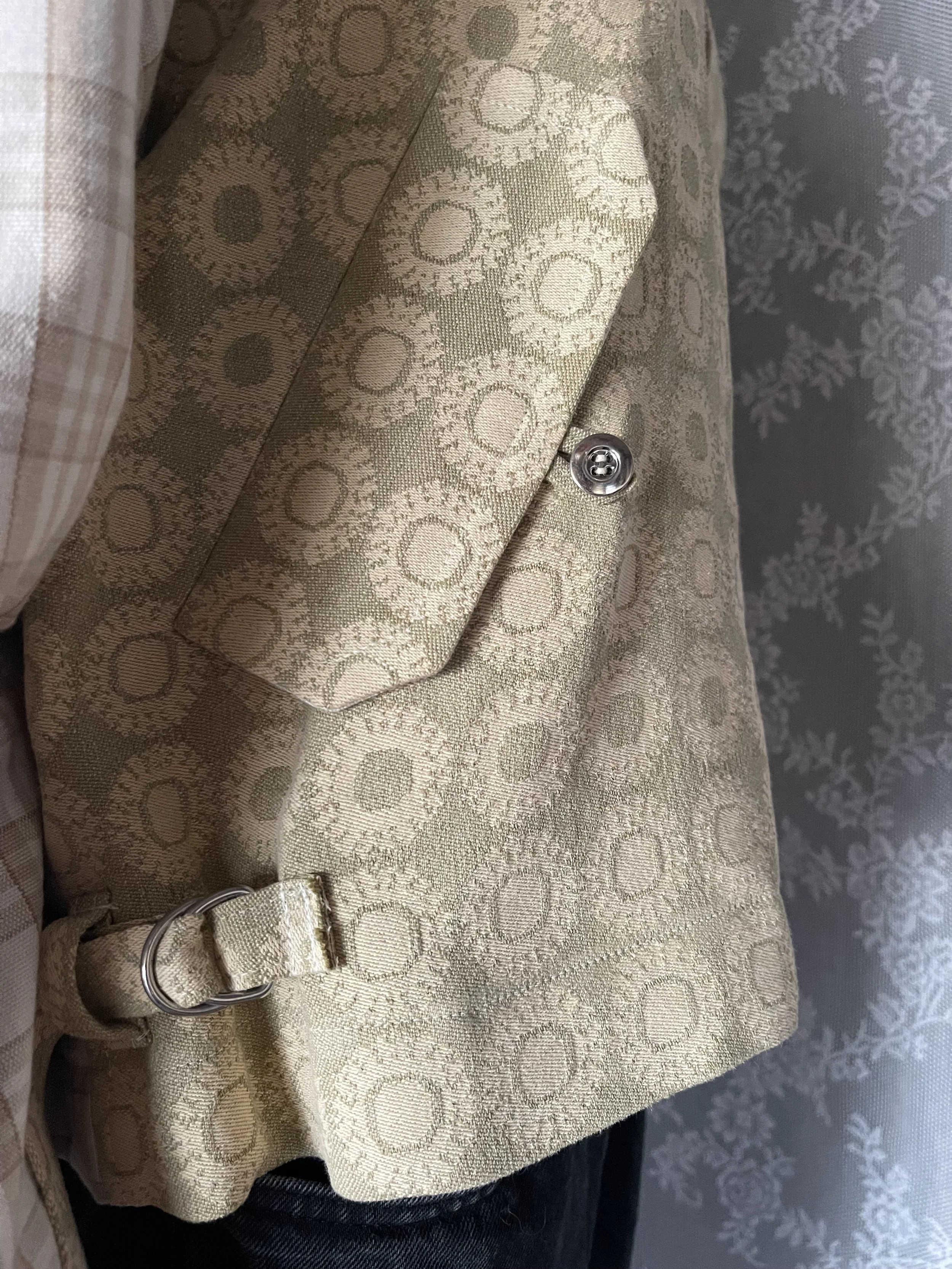 Close-up of a beige patterned blazer with a small silver button and a strap with a D-ring at the waist, and a glimpse of a plaid shirt sleeve on the left.