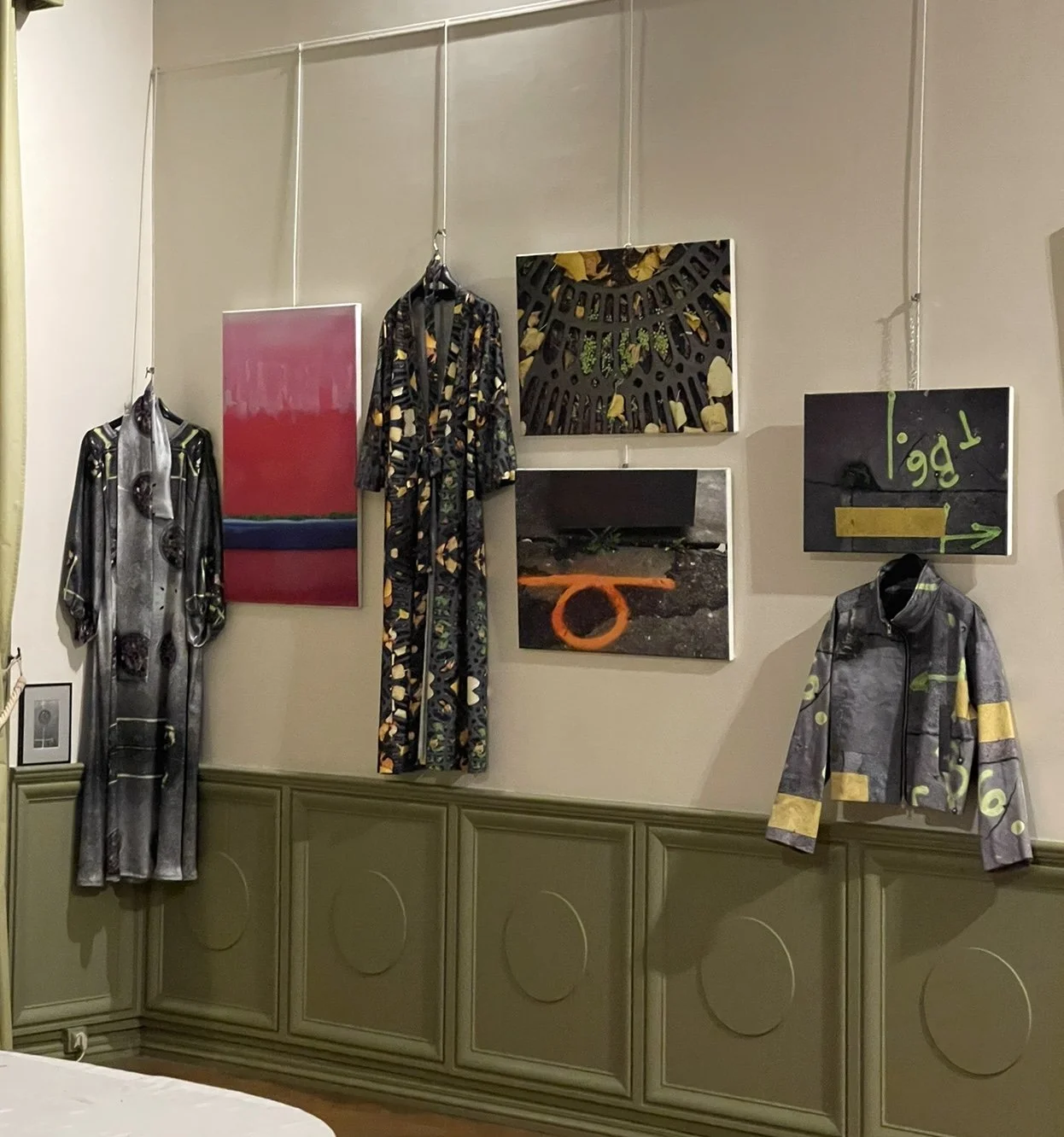 Gallery wall displaying two dresses and a jacket with abstract and patterned artwork, along with paintings including one with a road sign message, in an interior space.