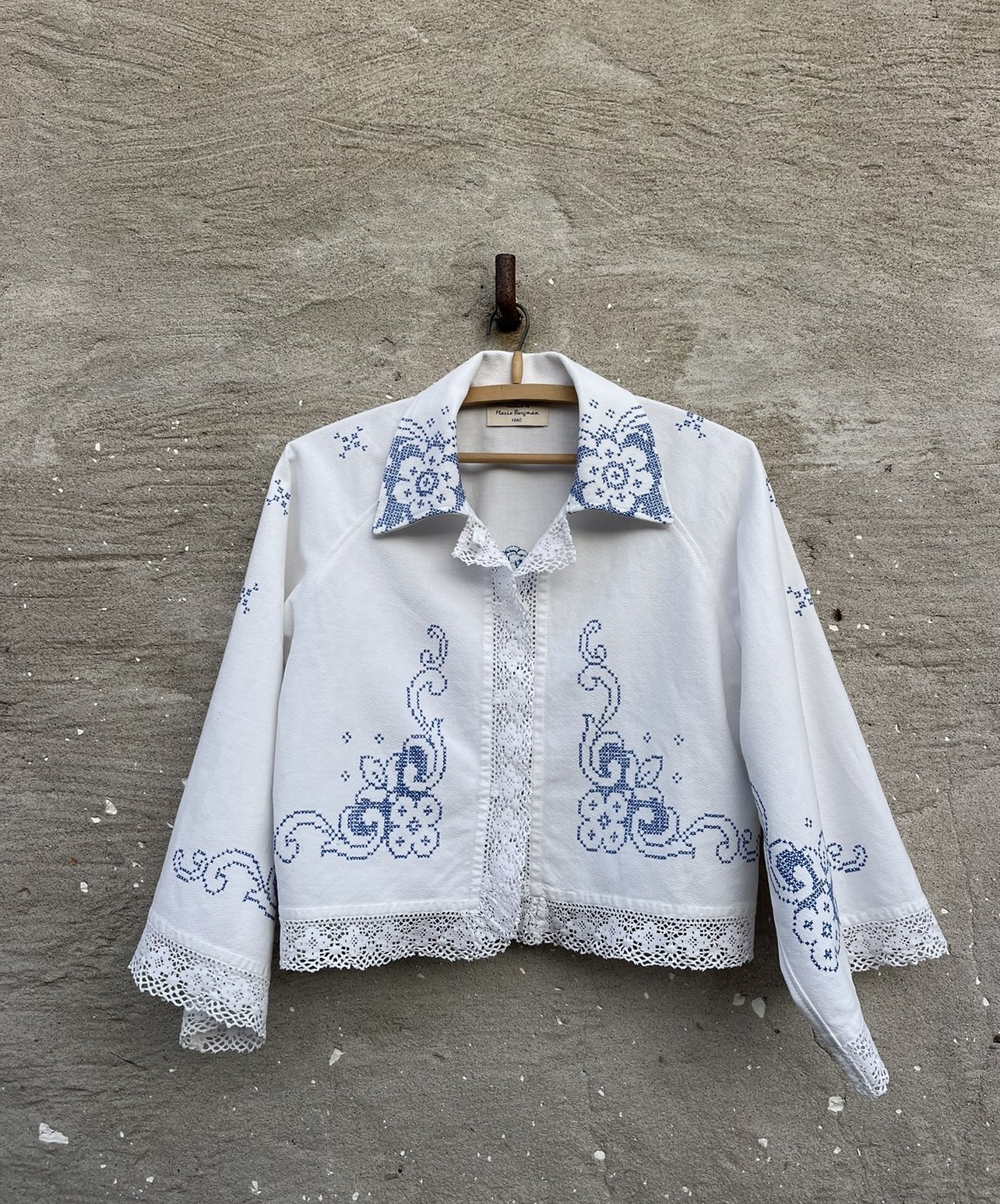 White embroidered jacket with blue floral patterns hanging on a wooden hanger against a concrete wall.