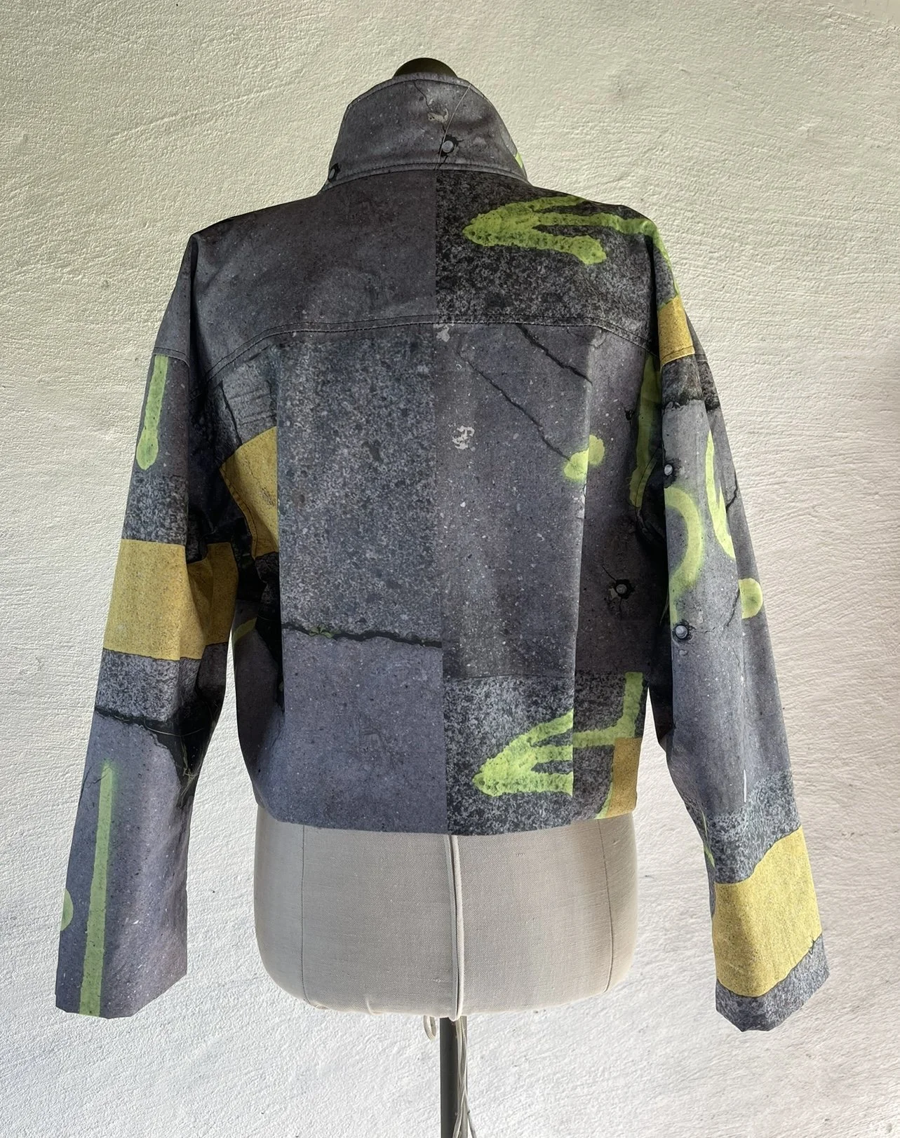 Back view of a jacket with an urban street art mural print featuring concrete textures and yellow-green accents, displayed on a mannequin against a light-colored textured wall.